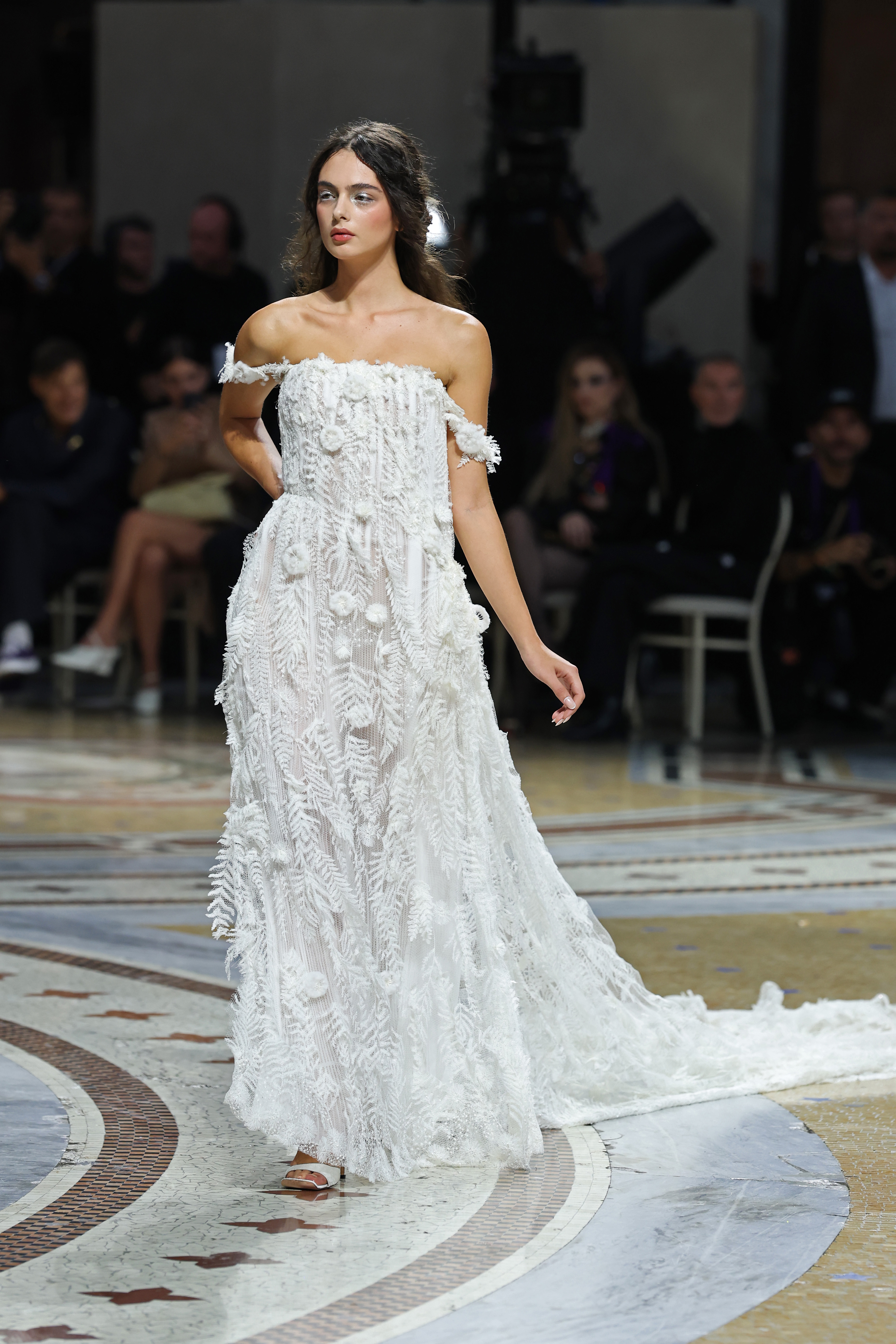 Mid-stride along the historic Galleria Vittorio Emanuele II corridor, Deva Cassel showcased the full, floor-length silhouette of her textured lace creation. The strapless design featured delicate off-the-shoulder accents and intricate botanical detailing down to a flowing, sheer-paneled hem as spectators admired the showcase. | Source: Getty Images