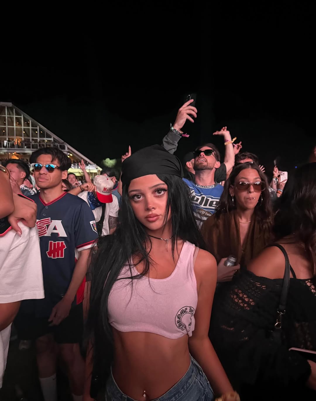 Harlow Winter Kate Madden poses in a white crop top and dark headscarf while attending Coachella 2026. Surrounded by festivalgoers at night, Kate faces the camera with a confident expression. | Source: Instagram/katemaddennn
