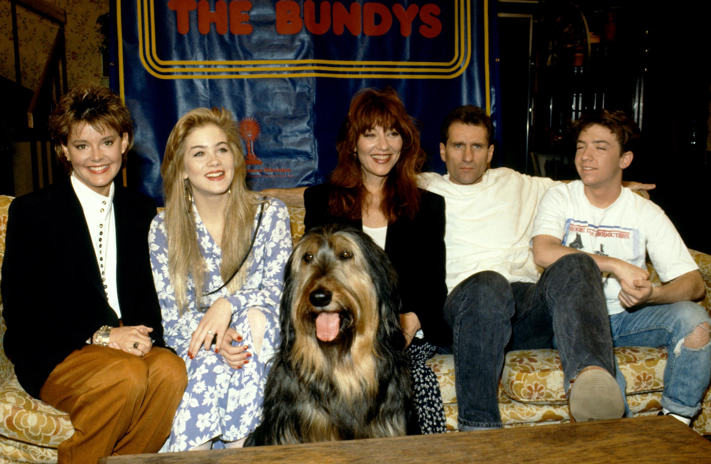 The cast of the American television sitcom "Married... with Children"—Amanda Bearse, Christina Applegate, Katey Sagal, Ed O'Neill, and David Faustino—crowded together on their floral living room sofa alongside their family dog, Buck, for a classic cast portrait in Los Angeles, California, circa 1990. Sitting beneath a custom "THE BUNDYS" banner, the group flashed cheerful smiles from the iconic set of the hit comedy series. | Source: Getty Images