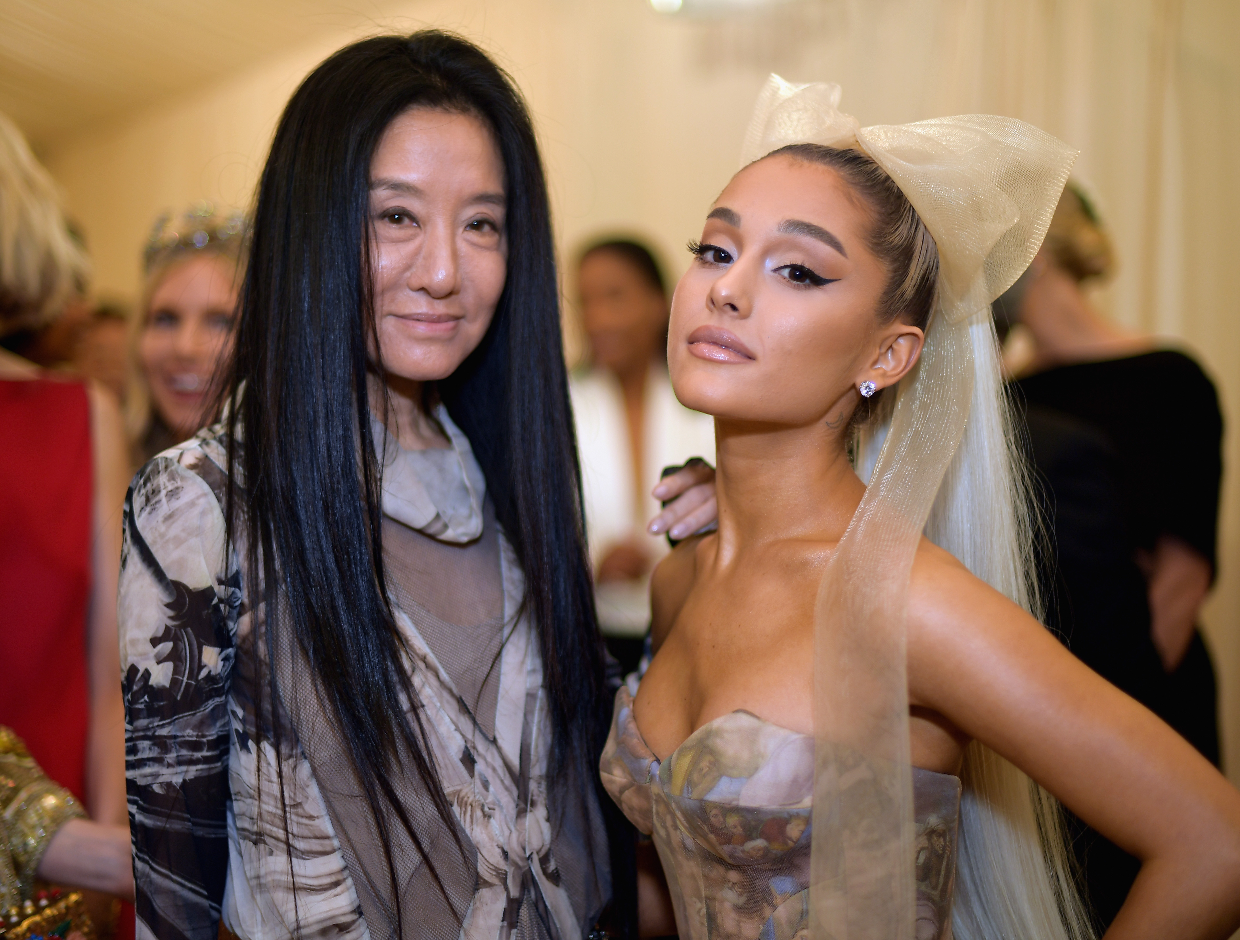 Vera Wang and Ariana Grande posed together inside The Metropolitan Museum of Art during the Heavenly Bodies: Fashion & The Catholic Imagination Costume Institute Gala on May 7, 2018, in New York City. Wang, who custom-designed the singer's Sistine Chapel-inspired ballgown, stood beside Grande wearing an airy printed sheer long-sleeve top over neutral layering, while the pop star showcased a dramatic winged eyeliner, platinum ponytail, and an oversized champagne tulle headbow. | Source: Getty Images
