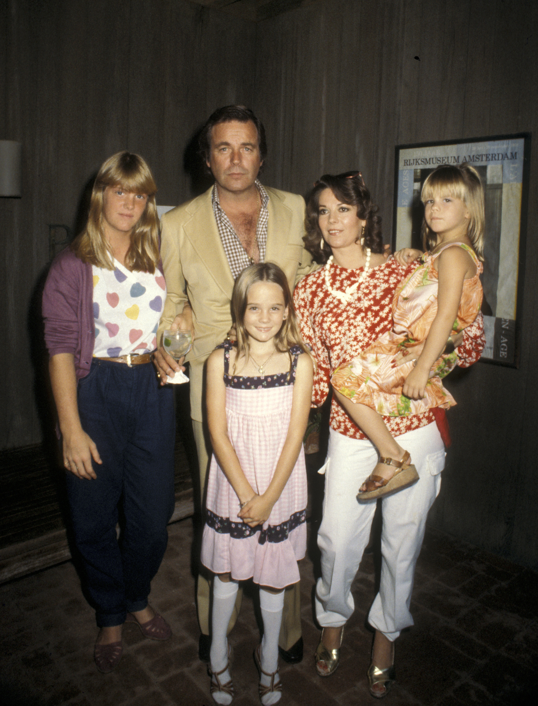 Robert Wagner and Natalie Wood pose with their daughters Katie Wagner, Natasha Gregson Wagner, and Courtney Wagner, circa 1981. Natalie holds Courtney in her arms while Natasha stands in front wearing a pink dress. Katie stands beside Robert, who holds a glass in one hand.
