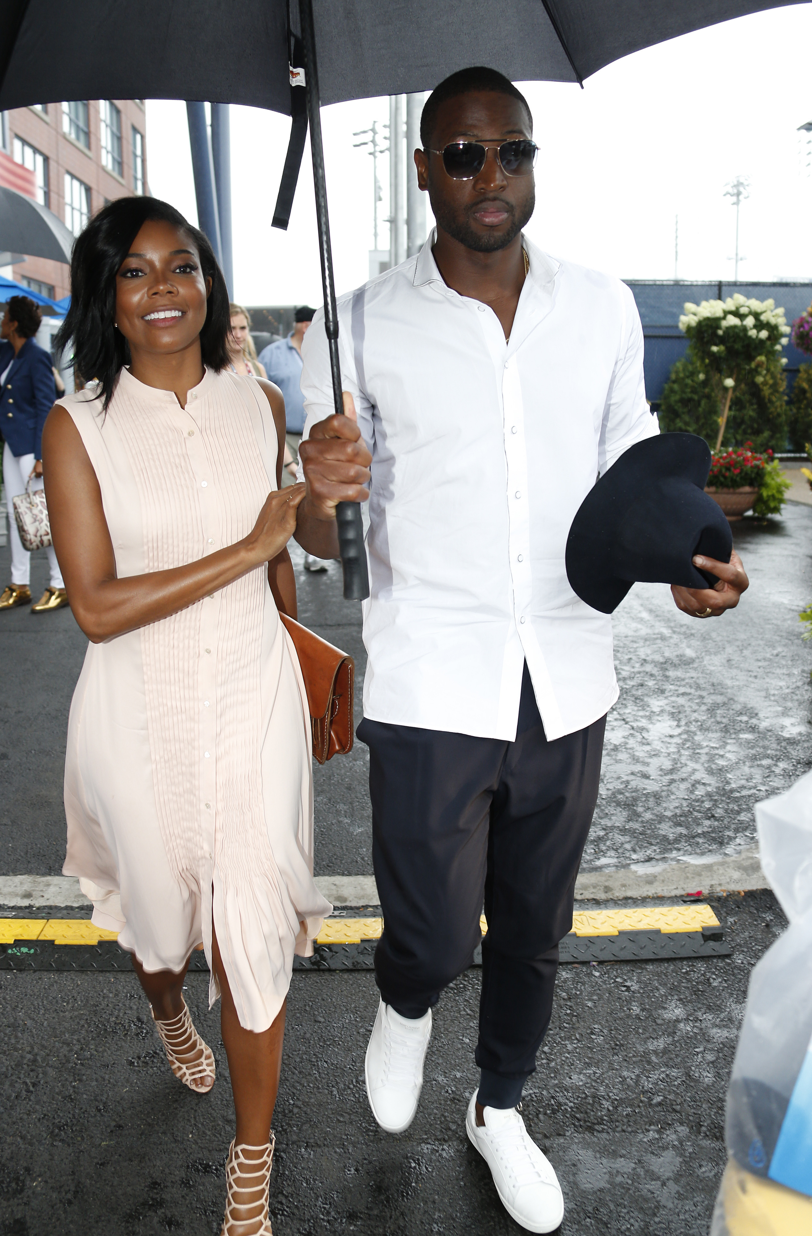 Gabrielle Union walks beside Dwyane Wade under a large umbrella on a rainy day. She wears a sleeveless, light beige dress with strappy heels and carries a small clutch, while he’s dressed in a crisp white shirt, dark trousers, sunglasses, and white sneakers, holding both the umbrella and a hat. | Source: Getty Images