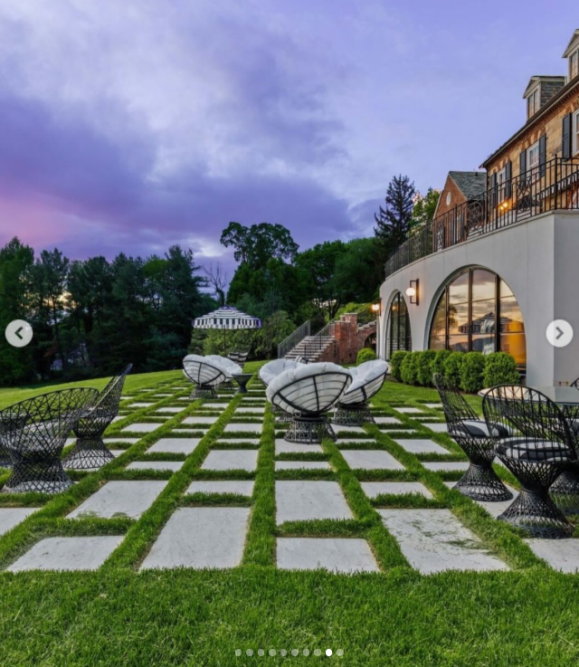 The outdoor entertaining area extends the living space onto the grounds, with large stone pavers, grass, and sculptural seating arranged beneath the terrace. Arched windows along the lower level connect this landscaped space directly to the mansion. | Source: Instagram/rogerperrygroup