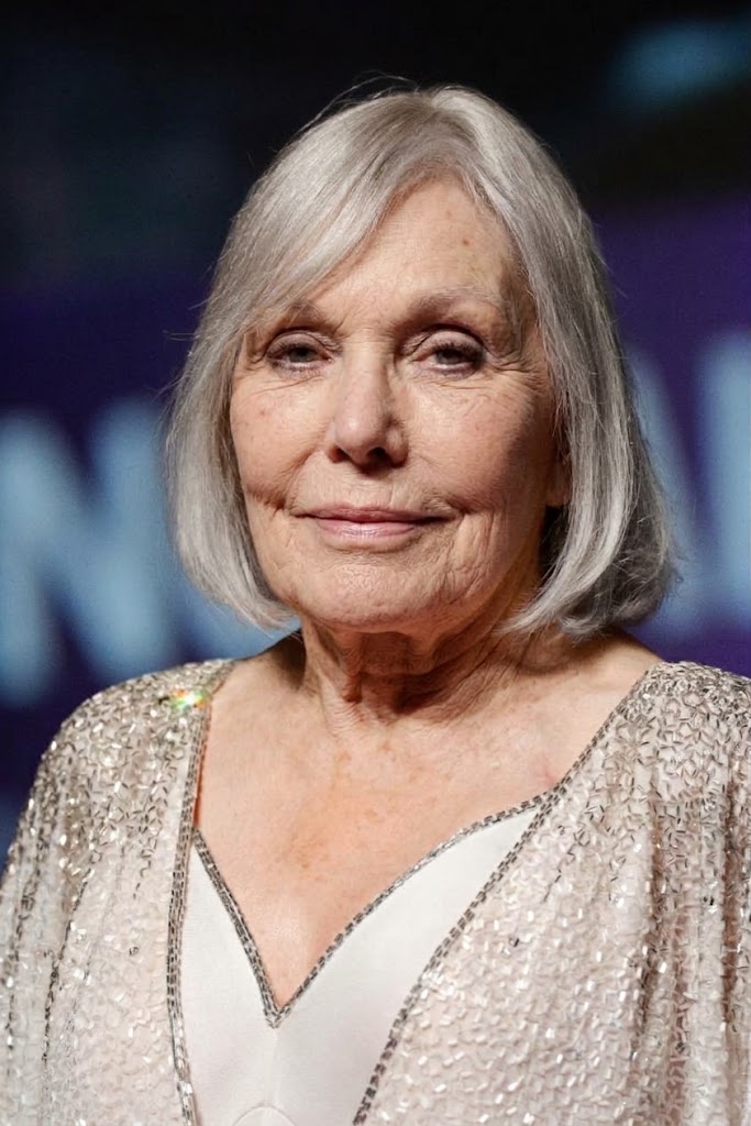 Kim Novak faces the camera with a composed expression, her silver-blonde bob complementing the glittering silver jacket and white dress against a softly blurred blue-purple background. | Source: Gemini