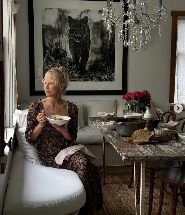 Dressed in a floral midi dress with a swept-up bun, Pamela Anderson relaxes on a white cushioned banquette in her rustic Arcady dining nook, holding a warm bowl of soup while gazing out the window. An ornate crystal chandelier hangs above a distressed wooden table set with fresh bread, roses, and steaming cookware, beneath a dramatic black-and-white wildlife print. She shared the tranquil mealtime scene with the message, "See the beauty in who you are, then share it. 🤍". | Source: Instagram/pamelaanderson