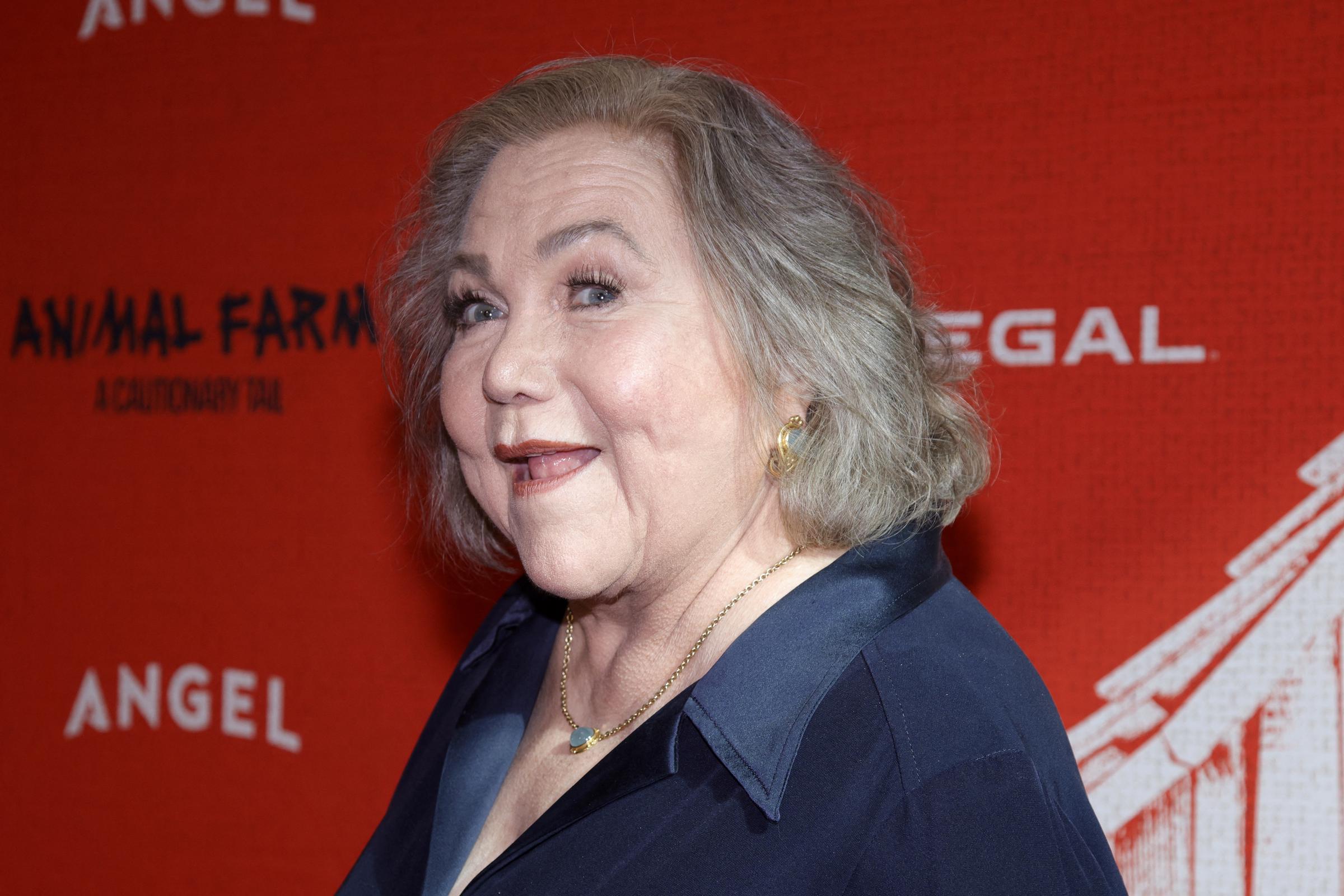 Kathleen Turner poses at the "Animal Farm" premiere, wearing a dark navy ensemble with gold jewelry against the event's bright red backdrop.