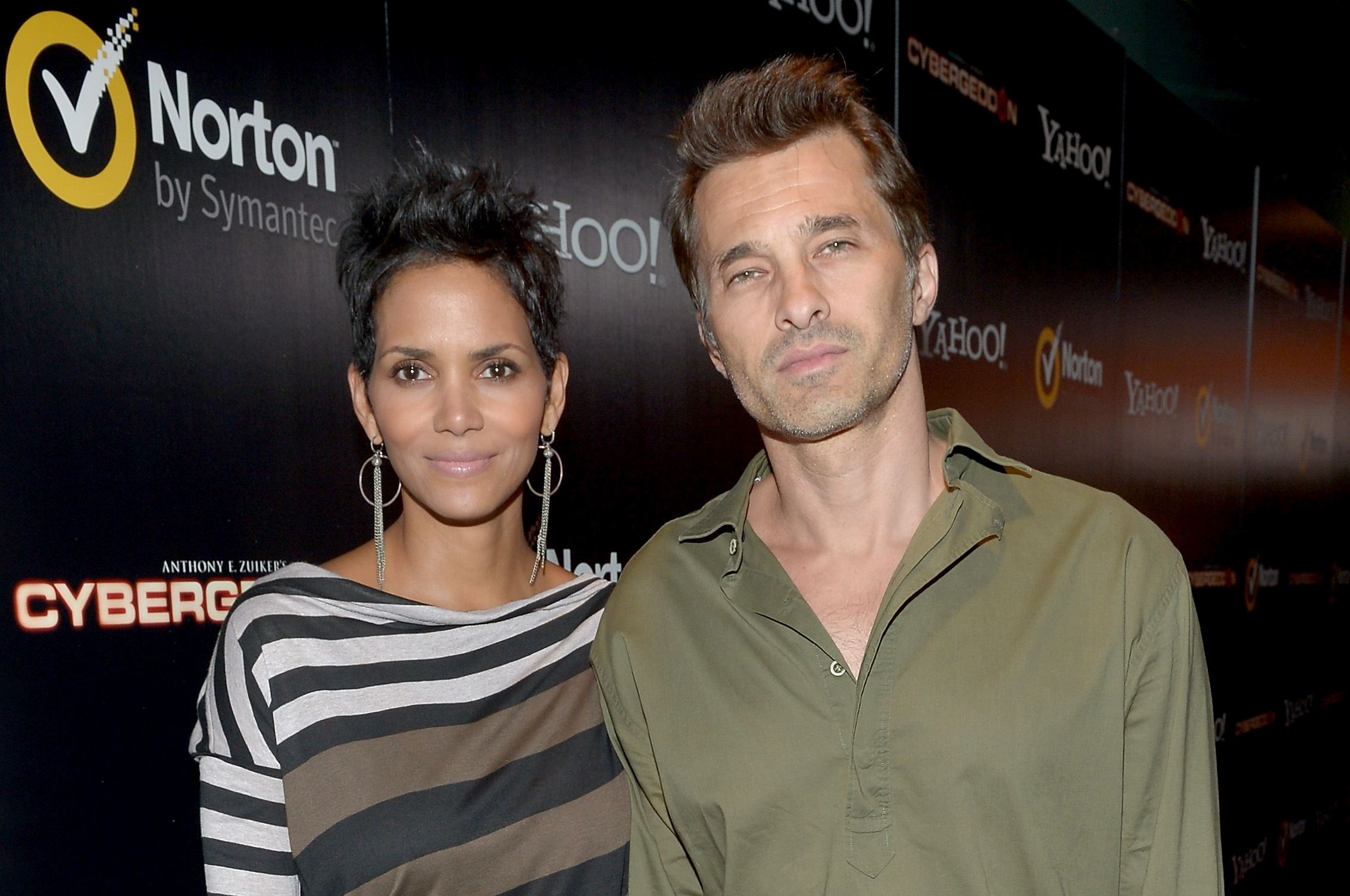 Actors Halle Berry and Olivier Martinez stood side by side on the black carpet during the premiere of "Cybergeddon" at the Pacific Design Center on September 24, 2012, in West Hollywood, California. Berry wore an asymmetrical boatneck top featuring bold horizontal stripes, styled with dangling fringe earrings and a textured, spiky pixie cut, while Martinez kept it relaxed in an open-collar olive green button-down shirt. | Source: Getty Images