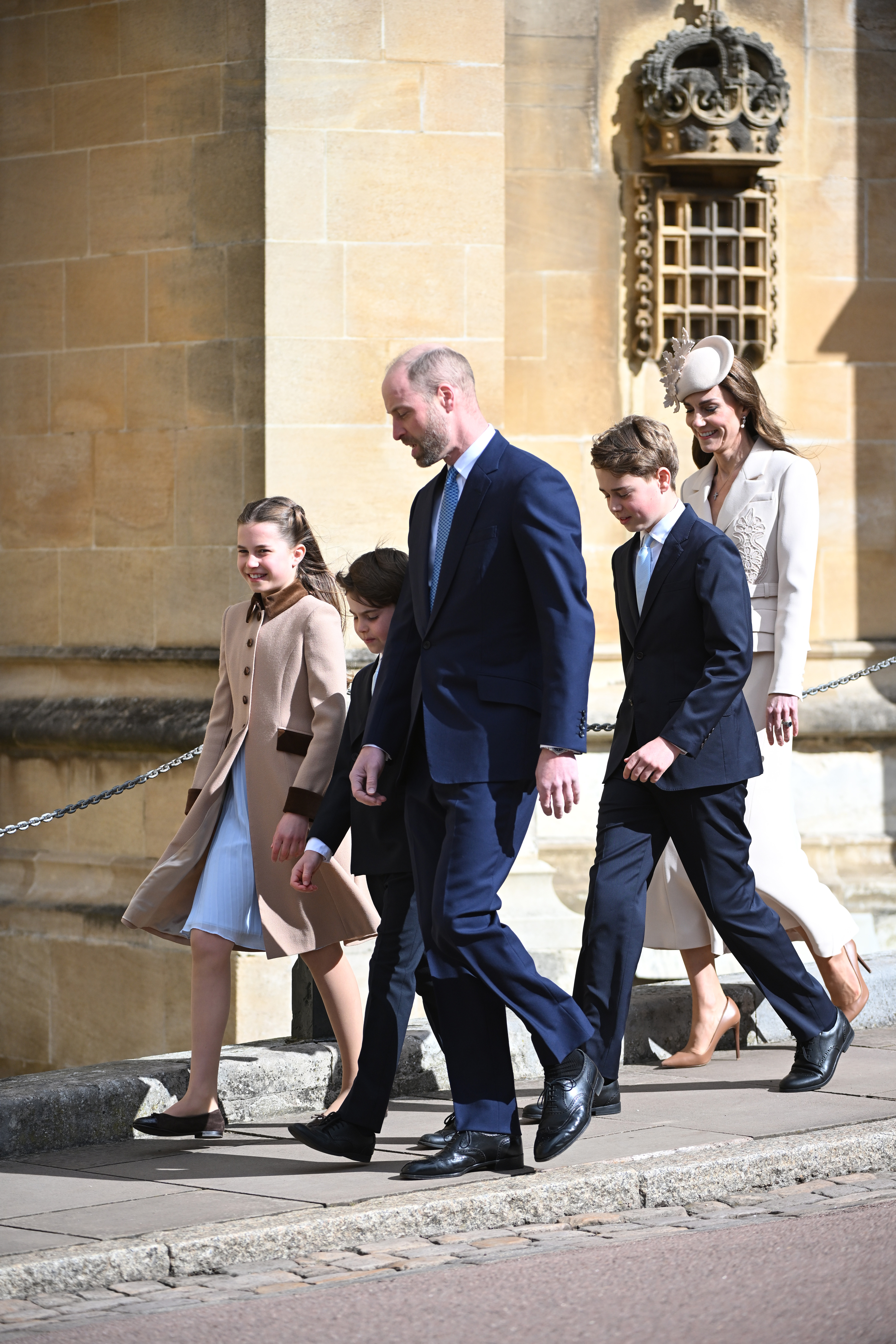 The family stepping up towards the chapel entrance, the ornate stone crown carved into the pillar above them. Princess Charlotte laughs as she walks on the left, Prince Louis and the Prince of Wales stride ahead in the centre, and Prince George walks alongside the Princess of Wales just behind them, the lace appliqué detail of her blazer catching the bright morning light. | Source: Getty Images