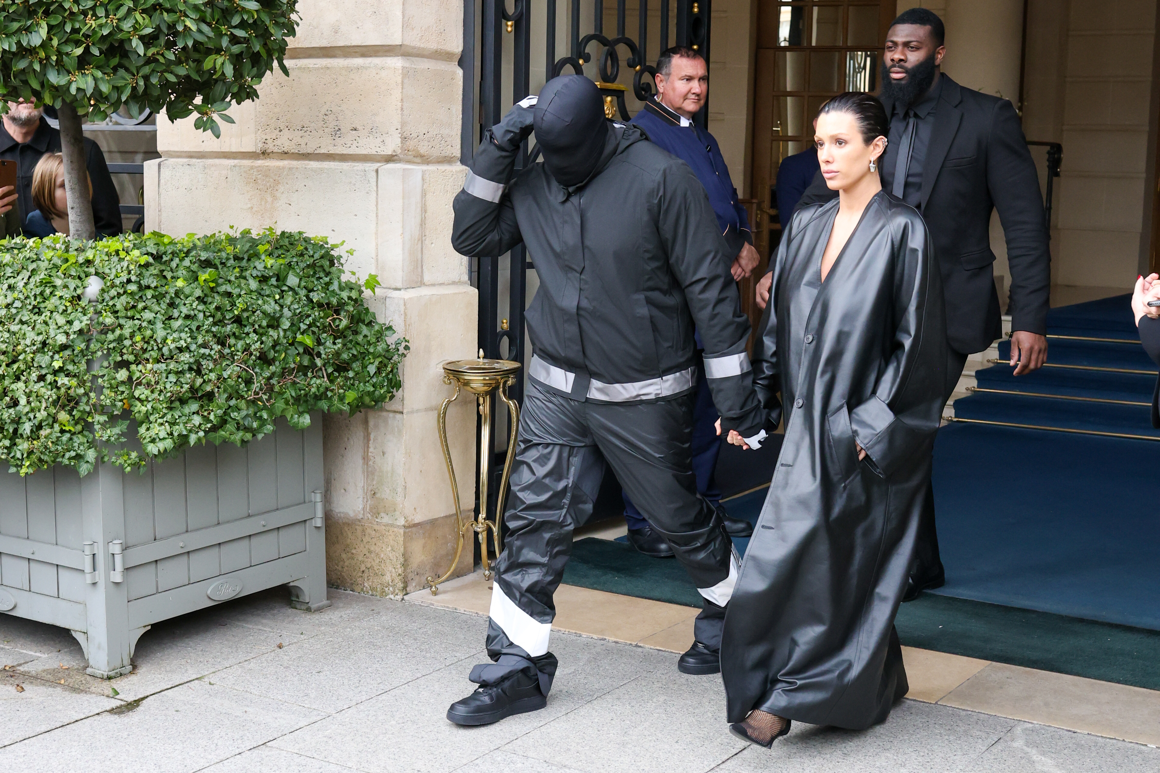 Kanye West and Bianca Censori stepped out in striking all-black silhouettes as they exited their Parisian hotel on February 28, 2024, in Paris, France. West remained completely incognito beneath a full-face black mask, utilitarian zip jacket, and reflective-banded cargo pants, guiding Censori by the hand as she swept past in a floor-length leather trench coat and slicked-back updo with security following closely behind. | Source: Getty Images