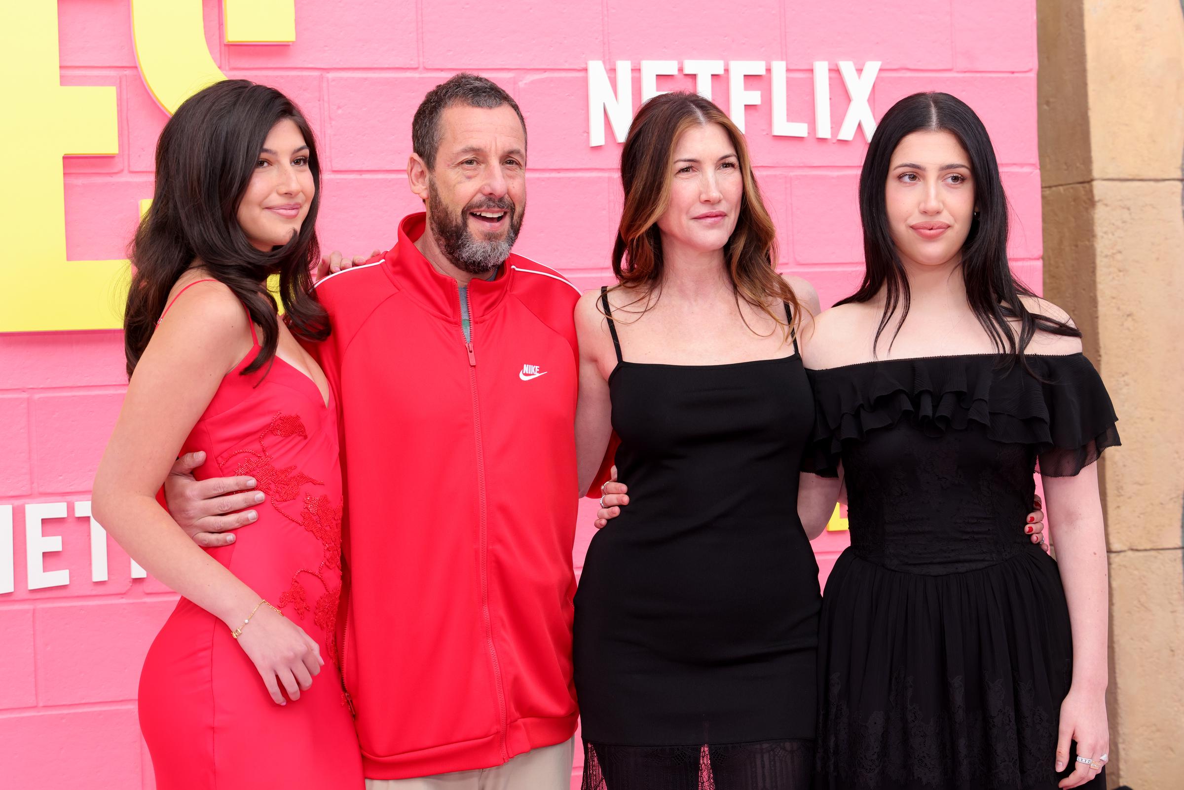 Sunny Sandler, Adam Sandler, Jackie Sandler, and Sadie Sandler pose closely, leaning into a coordinated red-and-black palette. Sunny stands out in bold red, while Jackie and Sadie keep it sleek in black beside Adam’s casual look.