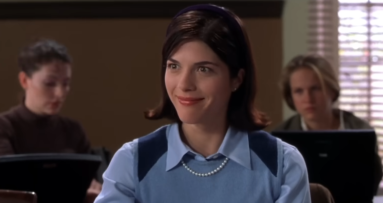 Selma Blair smiles smugly from her classroom desk as Vivian Kensington in a scene from the 2001 film "Legally Blonde". She wears a preppy light-blue collared shirt under a powder-blue sweater vest with navy shoulder accents, accessorized with a classic pearl strand necklace, stud earrings, and a dark headband holding back her flipped brunette bob. | Source: YouTube/Amazon MGM Studios
