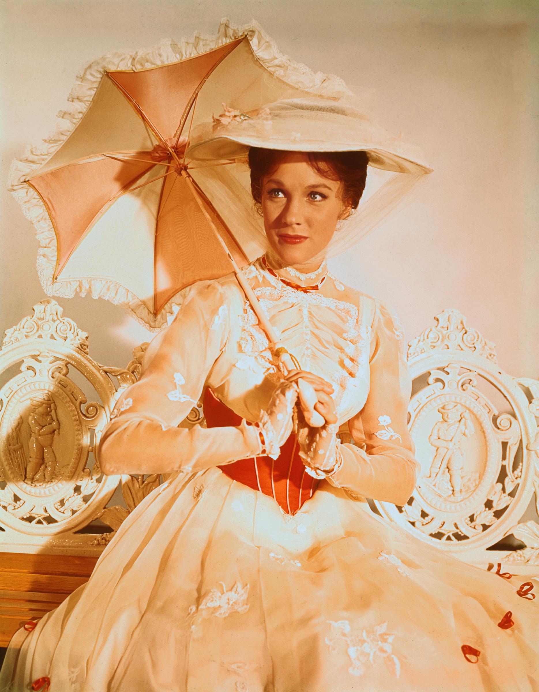 Julie Andrews epitomizes Edwardian elegance in a promotional portrait for the 1964 Disney musical classic "Mary Poppins." Dressed for the iconic "Jolly Holiday" sequence, the actress sits gracefully in an ornate white garden bench wearing the white ruffled organza dress accented with red ribbons, a wide-brimmed matching picture hat veiled with tulle, and holding her pink-and-white ruffled parasol.