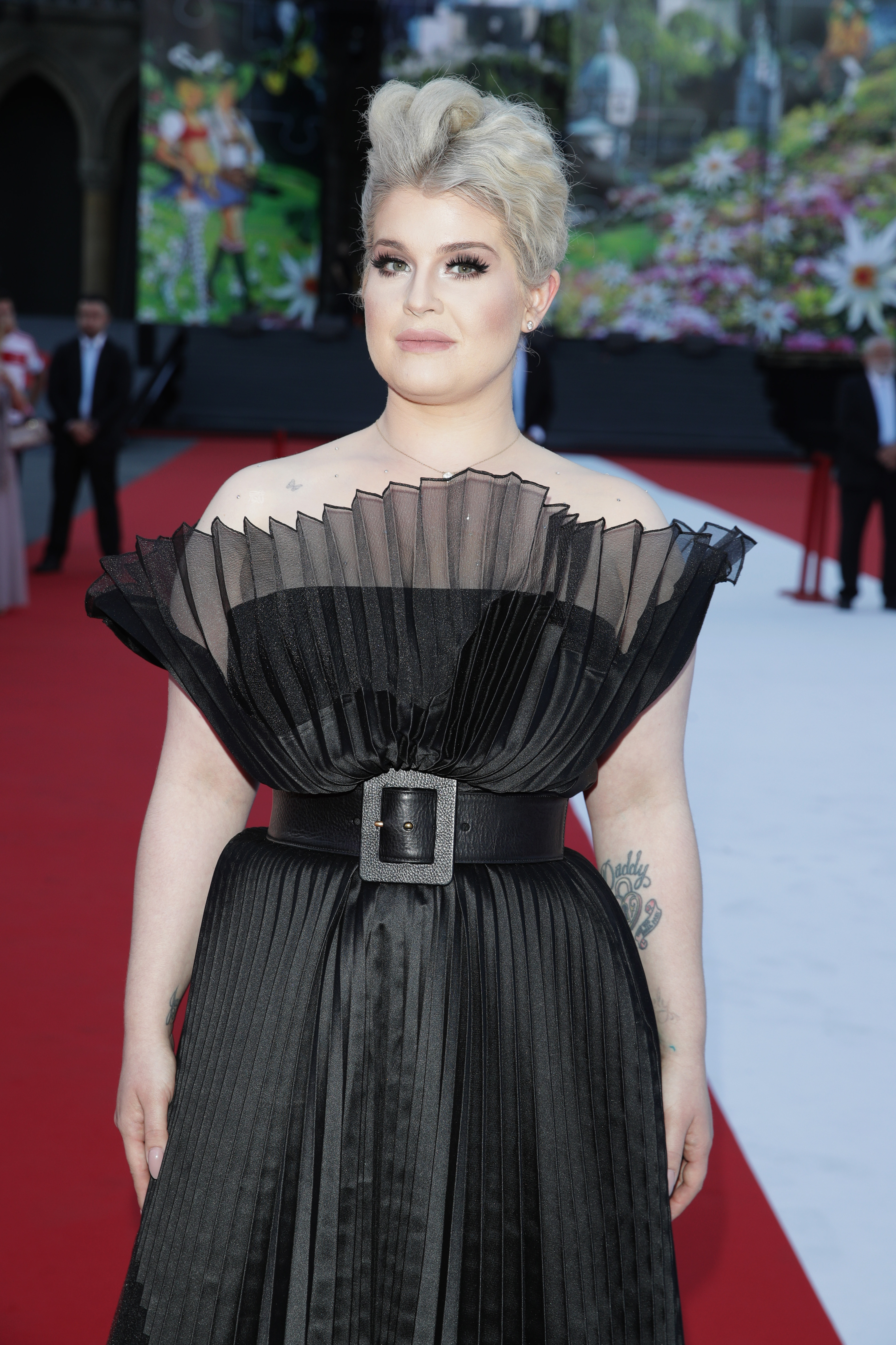 Kelly Osbourne arrives at the Life Ball 2018 at Vienna City Hall. The television personality wore a dramatic black pleated gown with an architectural ruffled neckline and a wide statement belt, complemented by a platinum-blonde updo and soft, glamorous makeup. | Source: Getty Images