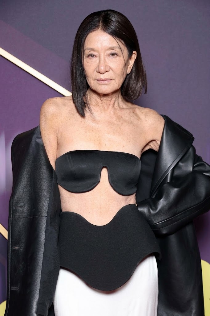 This AI-generated image envisions Vera Wang aging naturally while recreating her bold look from the 2025 EE BAFTA Film Awards red carpet in London. Reimagining the designer with a chic cropped bob, authentic fine lines, and sun-kissed mature skin texture, the digital concept portrays her in the headline-making sculptural black bandeau top, matching curved peplum waist, draped black leather outerwear, and white floor-length skirt. | Source: Gemini