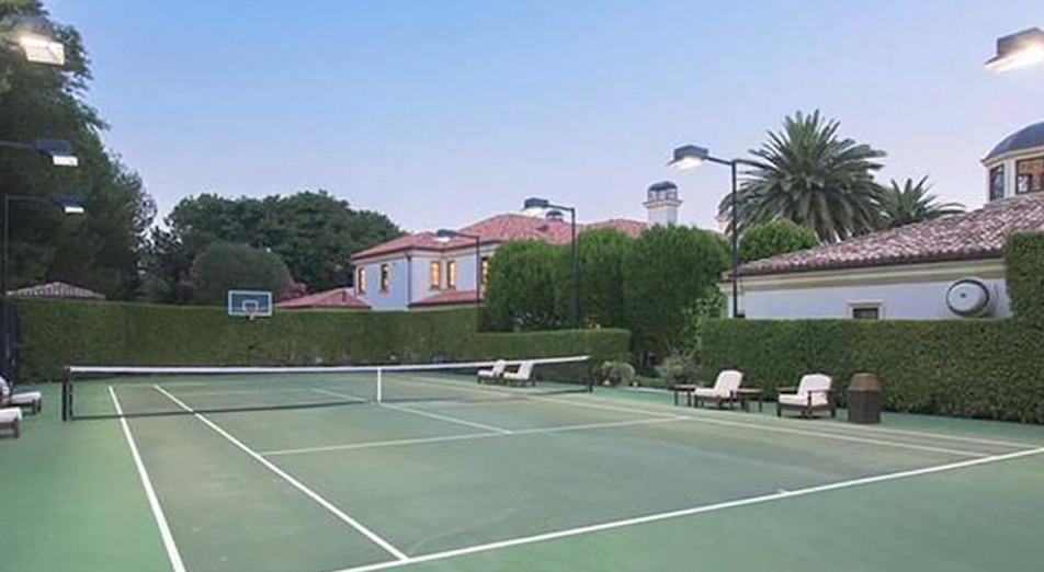 A private, championship-style regulation tennis court features nighttime floodlights, an integrated basketball hoop, and manicured privacy hedges that screen the neighboring estate grounds. | Source: YouTube/Taste of Country