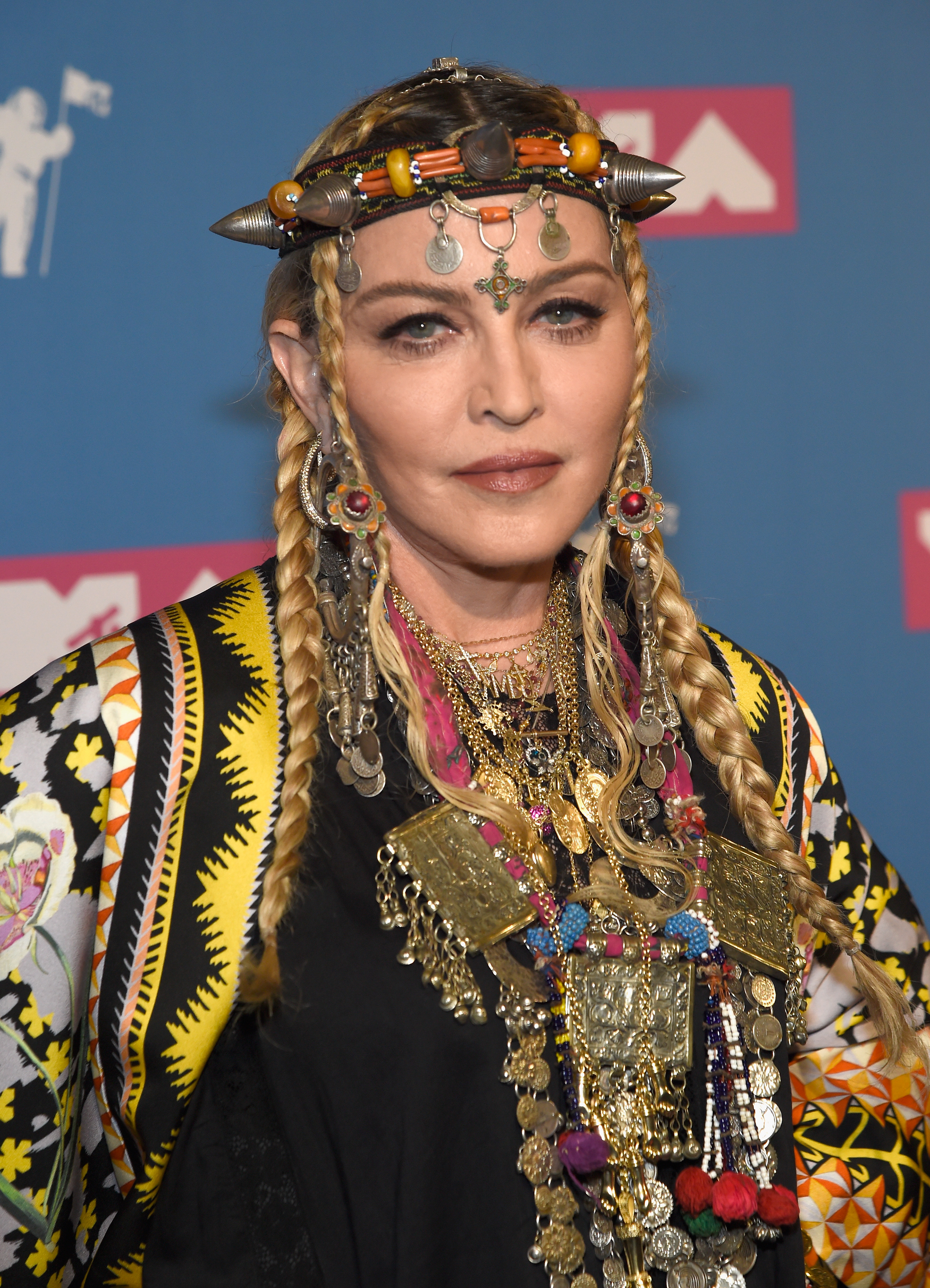 Madonna faces the camera in long blond braids, an ornate headpiece, oversized earrings, layered jewelry and a brightly patterned outfit. | Source: Chat GPT