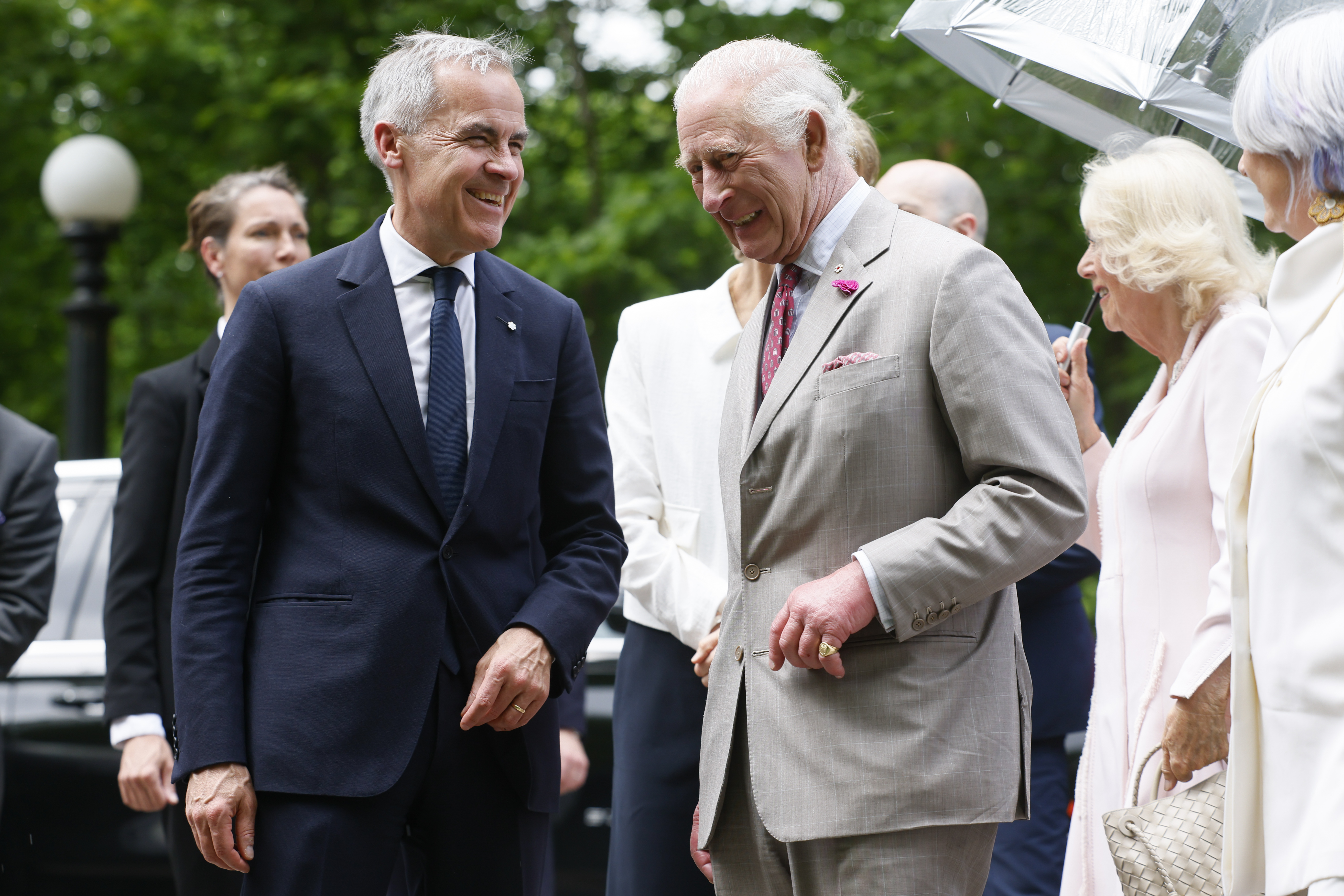 King Charles III and Prime Minister Mark Carney share a light moment, both laughing as their conversation brings a spark of warmth to the official welcome. In the background, Queen Camilla holds an open umbrella as she interacts with other officials.