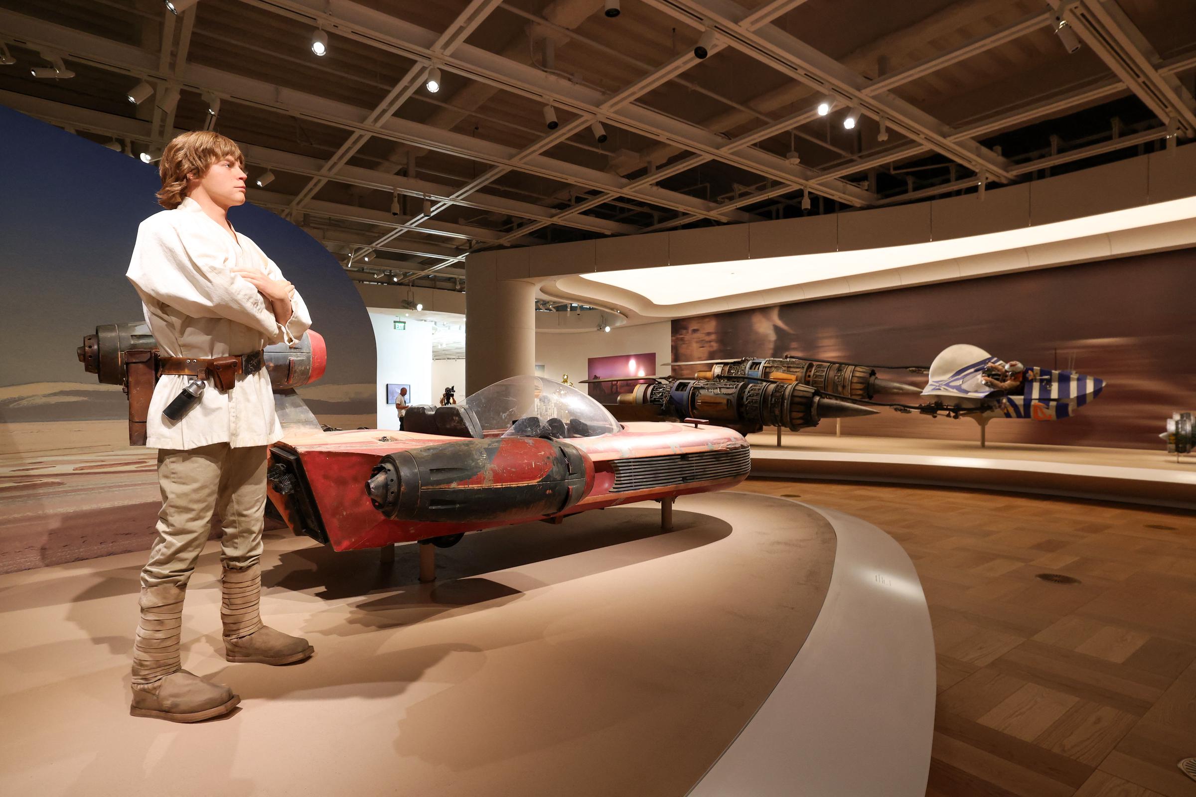 A "Star Wars" gallery brings familiar characters and vehicles into the museum setting, with a figure of Luke Skywalker positioned beside a red landspeeder. Additional vehicles appear throughout the exhibition space behind him. | Source: Getty Images