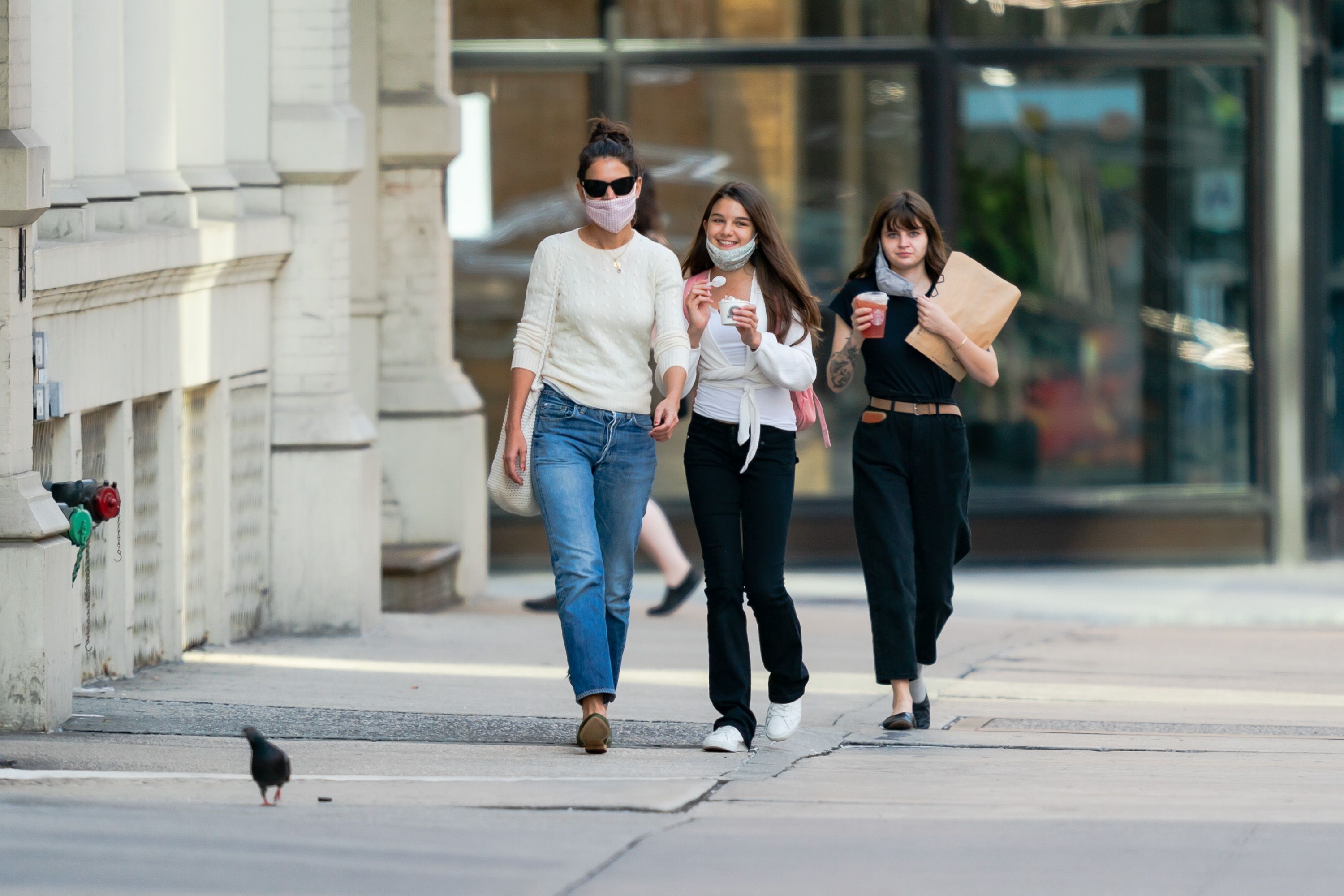 In this candid street scene, a grown Suri Cruise walks alongside her mother, Katie Holmes, and another unidentified individual. All three wear masks amid the pandemic, blending into the city's cautious rhythm. Suri, however, has slightly lowered her mask, revealing a warm, fleeting smile.