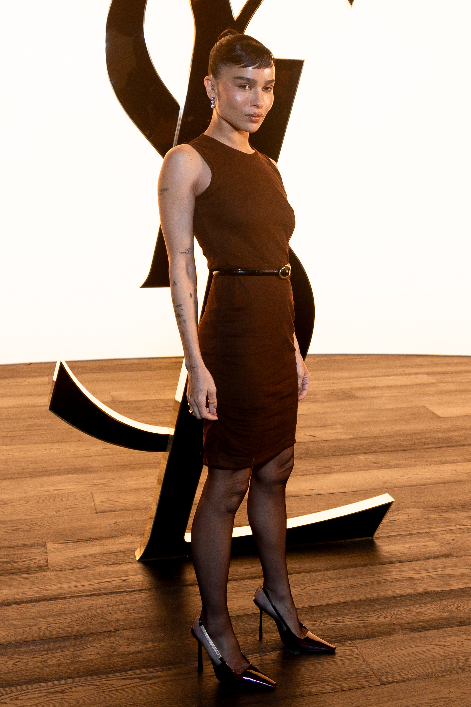 Zoë Kravitz attended the Saint Laurent Womenswear Fall/Winter 2024–2025 show during Paris Fashion Week on February 27. She wore a sleeveless brown knee-length dress cinched with a slim belt, paired with sheer tights, black pointed heels, and a sleek updo with side-swept bangs.