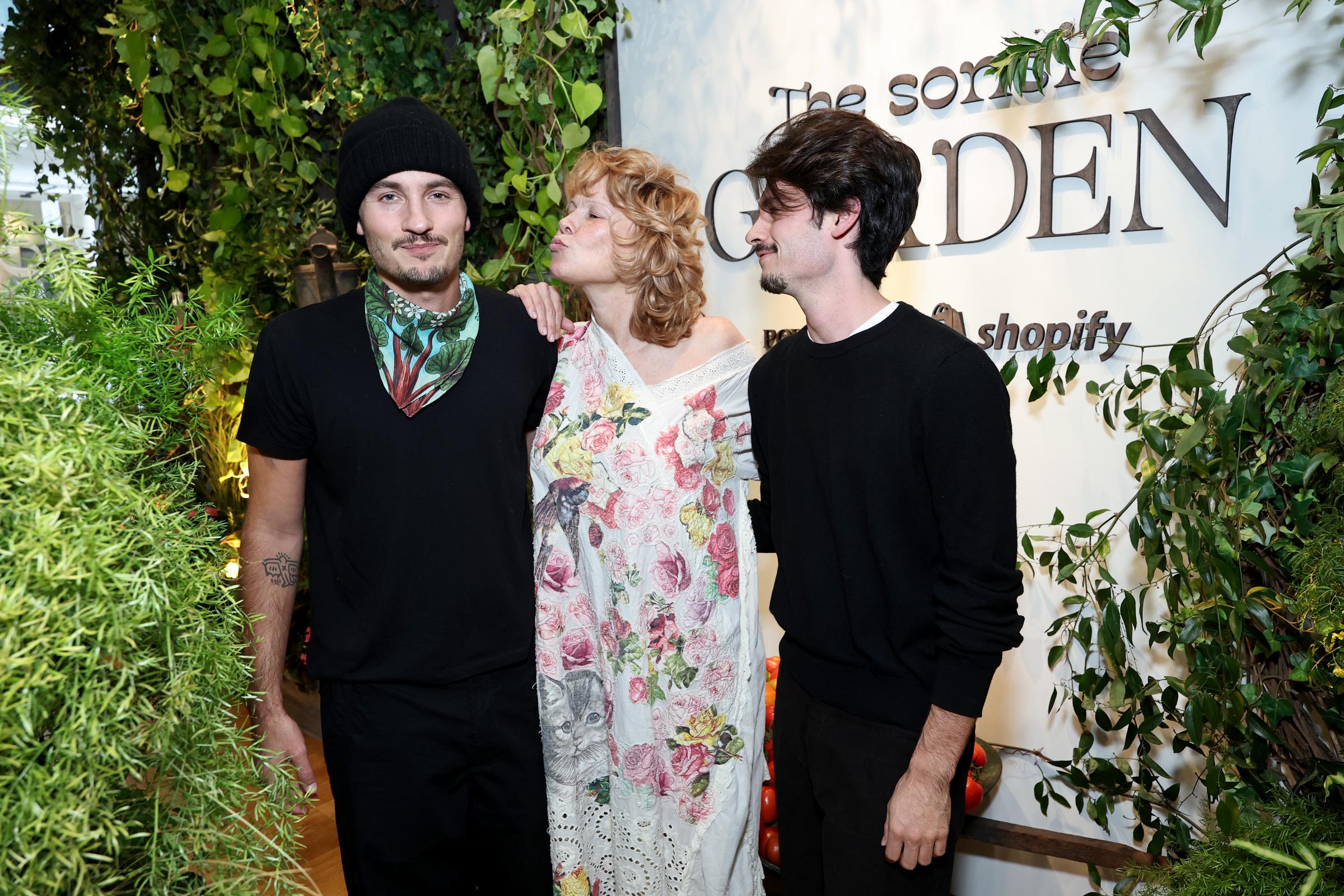 Brandon, Pamela, and Dylan pose for photos. Pamela stands between her sons wearing a white floral dress, leaning in to kiss Brandon's cheek.