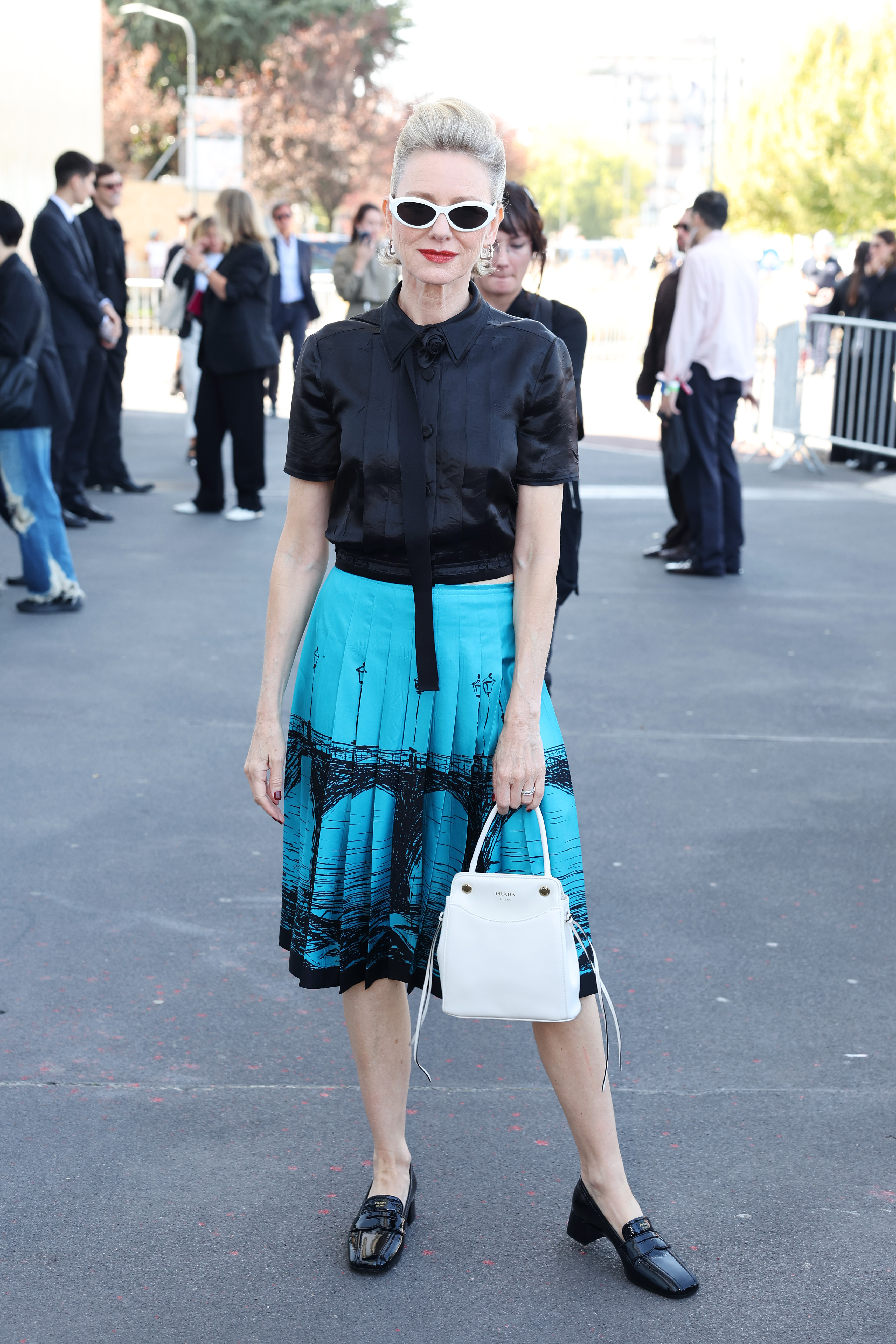 Watts faces photographers in white cat-eye sunglasses while holding a structured white handbag against her turquoise-and-black look. | Source: Getty Images