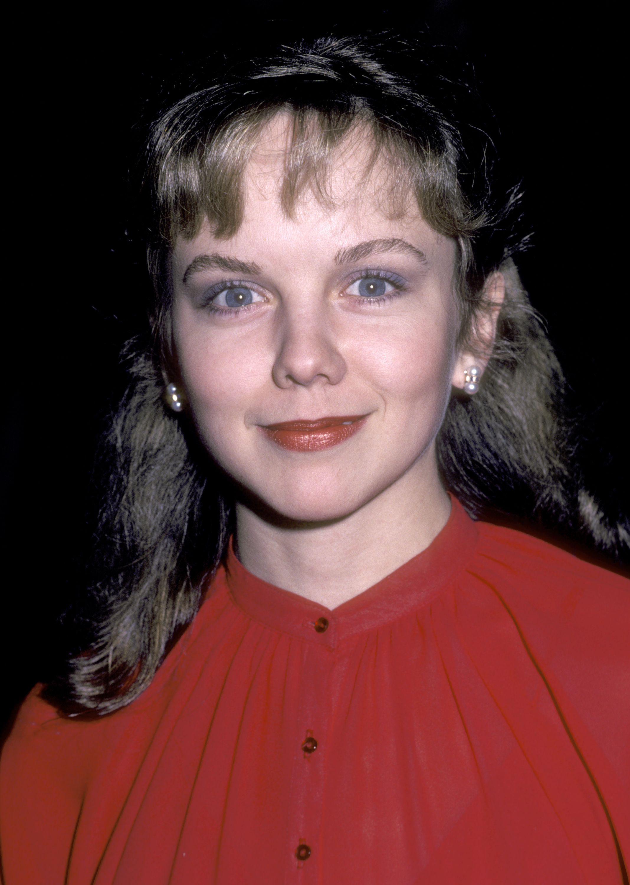 Attending the 18th Annual Publicists Guild of America Awards at the Beverly Hilton Hotel on March 27, 1981, 25-year-old Linda Purl paired an innocent, doll-like gaze with elevated evening glamour. Smoky lavender eyeshadow and a glossy brick-red pout framed her soft, cherubic features, while a finely pleated red high-collar blouse, pearl cluster earrings, and textured brunette hair with feathered bangs completed the poised early-1980s look.