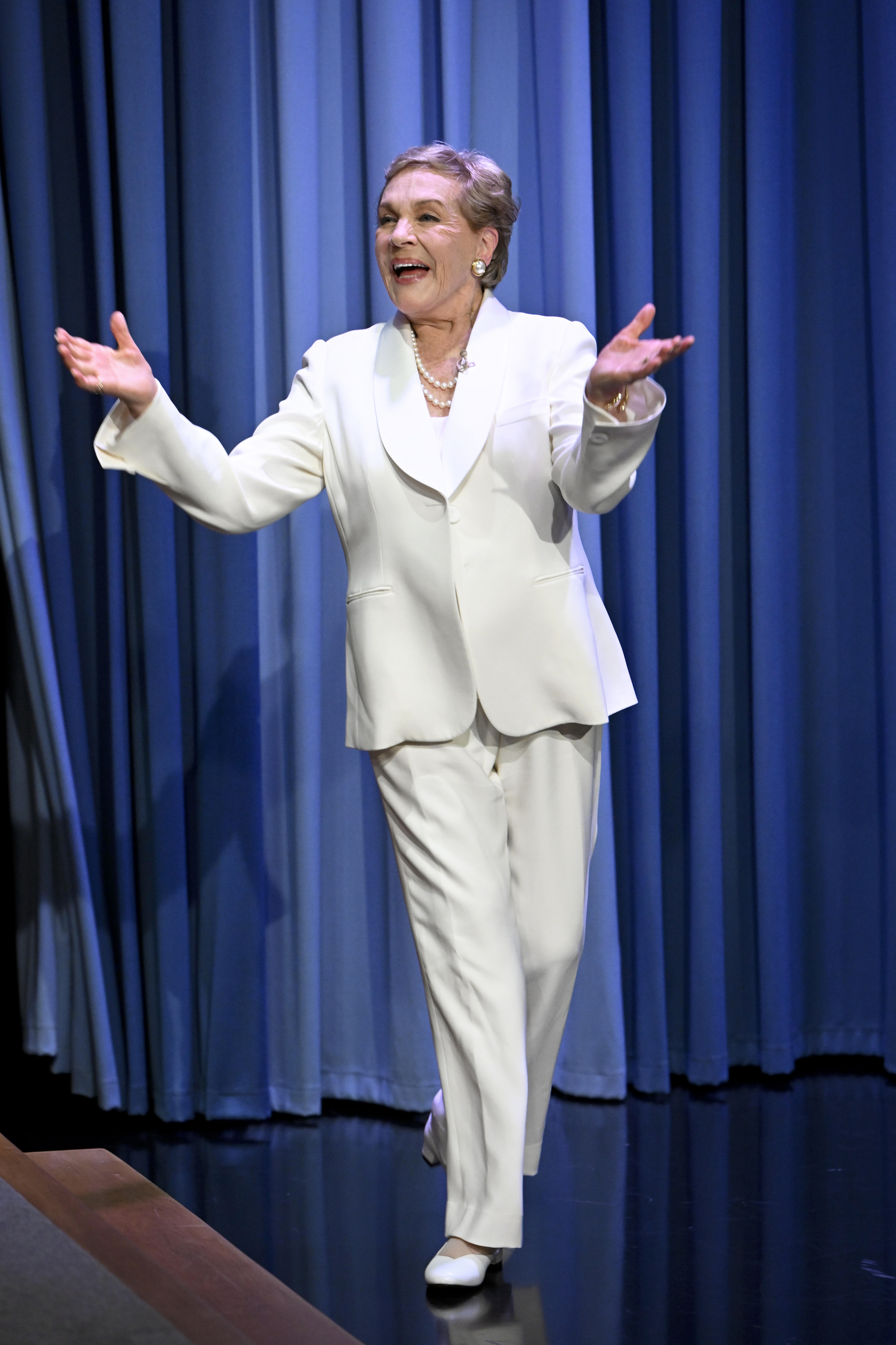 Julie Andrews arrives on stage with arms outstretched and a joyful smile during an appearance on "The Tonight Show Starring Jimmy Fallon" on June 24, 2022. Exuding effortless sophistication against the studio's iconic blue curtains, the entertainment legend wears an all-white tailored pantsuit with a rounded lapel blazer and white flats, accessorized with a layered pearl necklace, pearl cluster earrings, and a delicate bracelet.