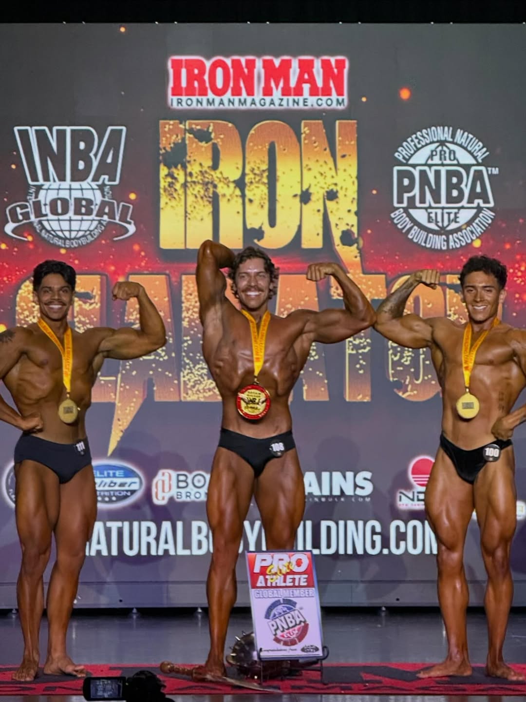 Joseph Baena hits a front double biceps pose between two competitors during a judging round, emphasizing symmetry and control. | Source: Instagram/joebaena