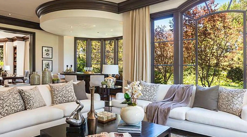 Sprawling white sofas accented with textured taupe pillows anchor the light-filled lounge, set against grand arched windows framing mature trees, dark architectural molding, and an adjacent curved wet bar positioned beneath a circular ceiling soffit. | Source: YouTube/Taste of Country