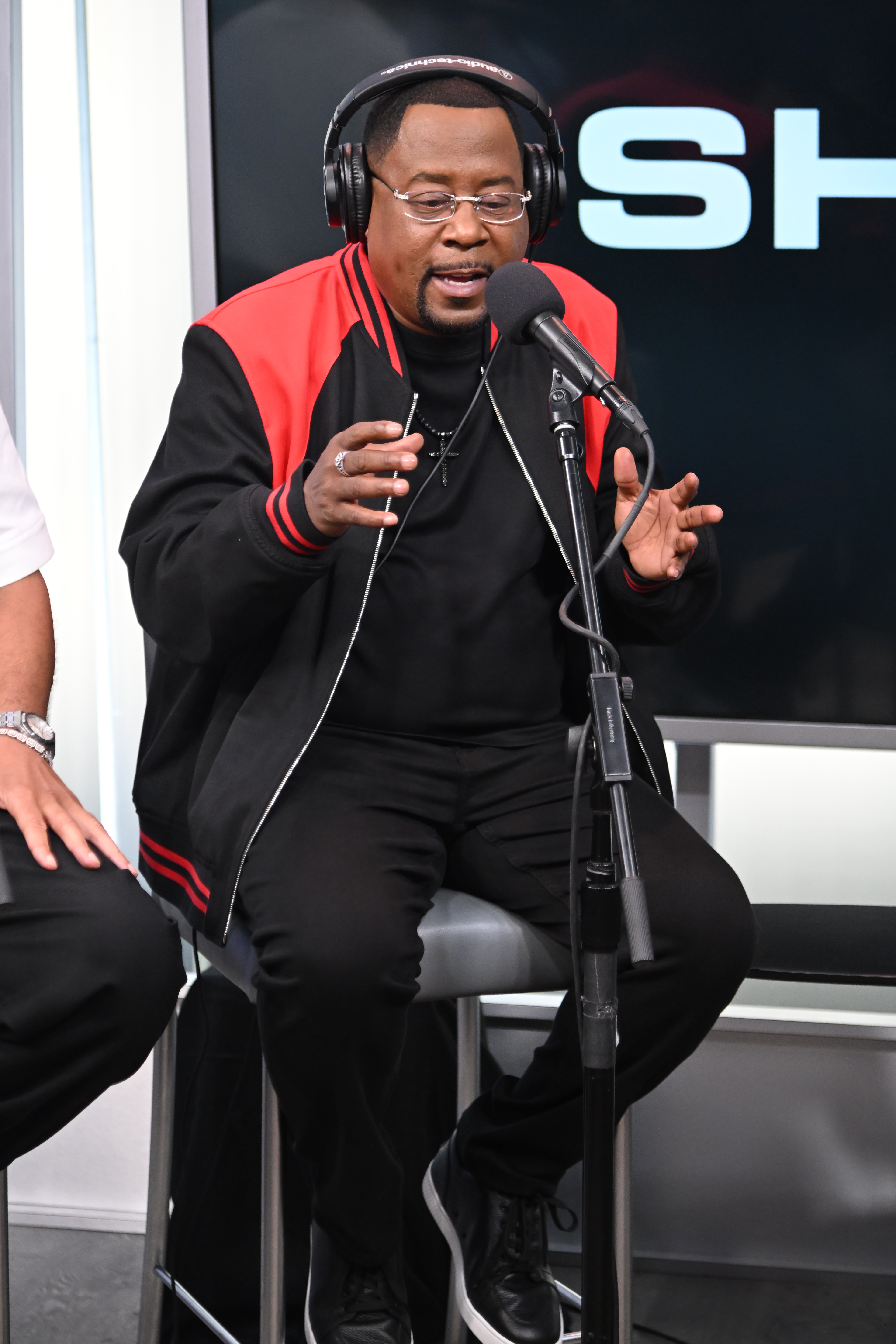 Seated at SiriusXM Studios, Martin Lawrence speaks into a microphone during a Town Hall event. Wearing headphones and a black outfit with red accents, he gestures with his hands mid-conversation, appearing engaged and expressive.