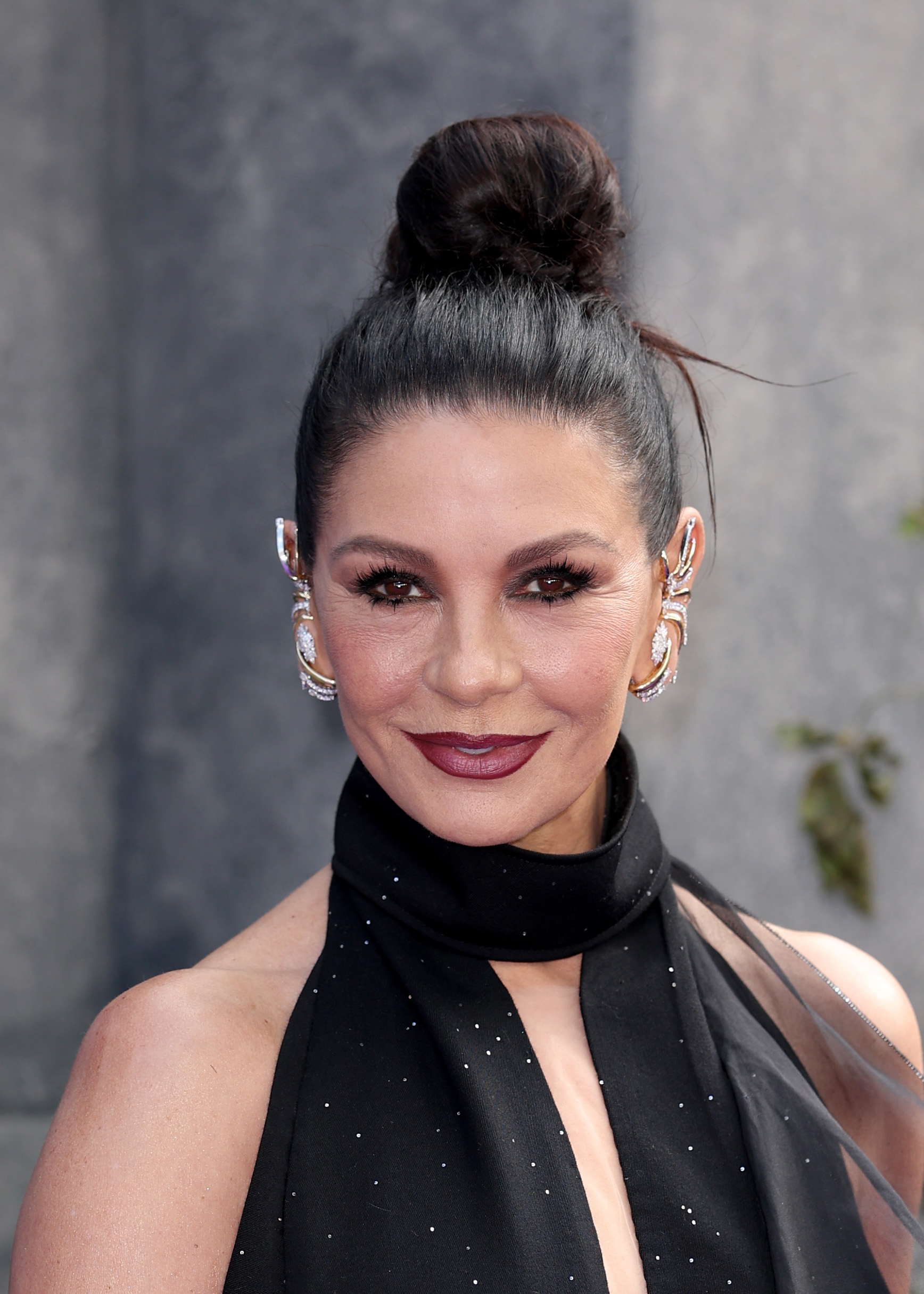 Catherine Zeta-Jones smiles on the purple carpet at the global premiere for Season 2, Part 1 of "Wednesday" at Central Hall, Westminster on July 30, 2025, in London, England. Channeling gothic elegance fitting for her role as Morticia Addams, the actress wears an embellished halter neck black gown with subtle crystal dusting, her dark hair swept up into a sleek high bun that showcases dramatic diamond-and-gold ear cuffs, winged eye makeup, and a deep berry-wine lip.
