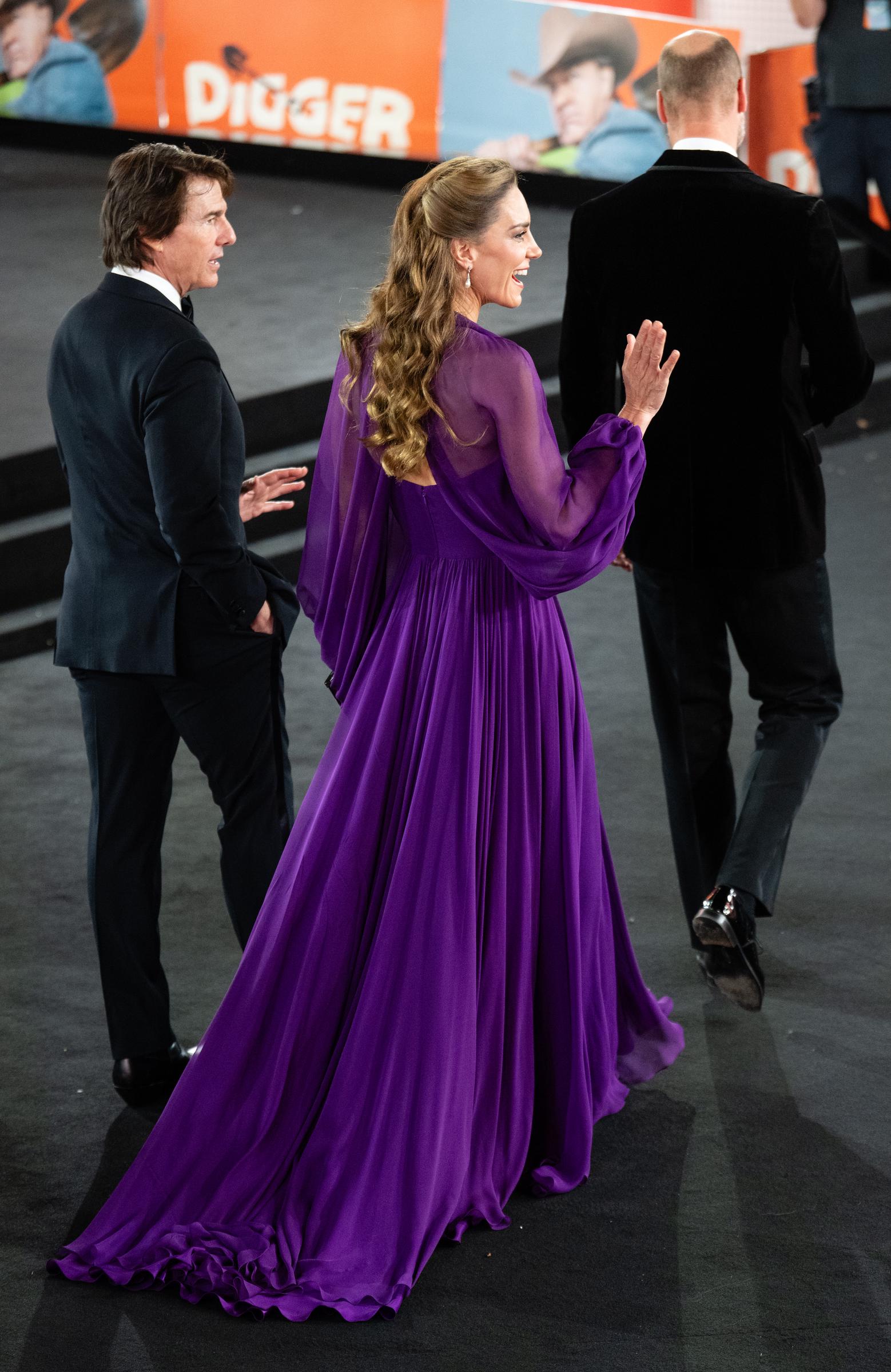 Catherine is photographed with Tom and William during the premiere, with the back of her gown visible as she turns. The floor-length design features sheer sleeves, draped fabric, and a long train.
