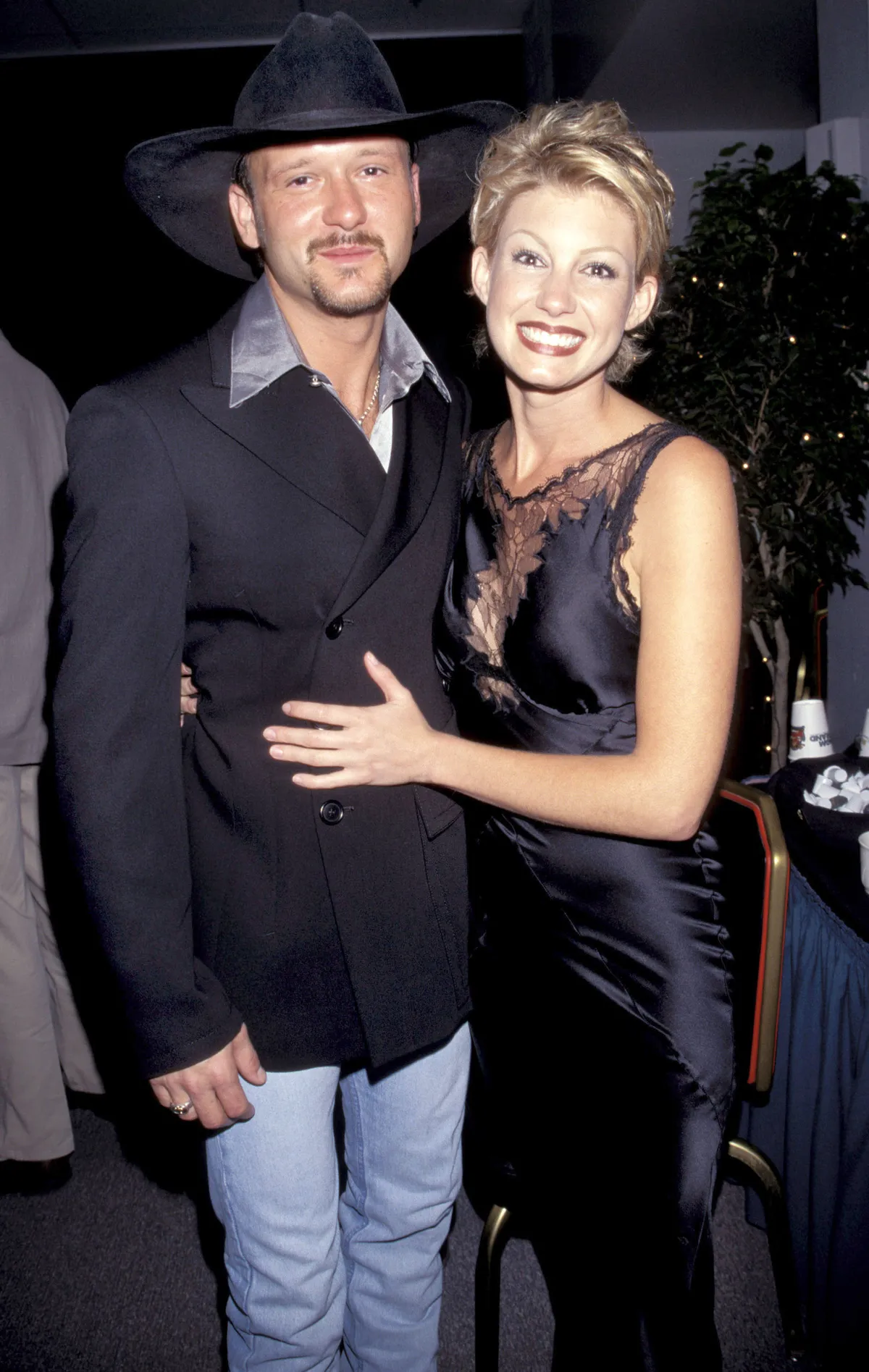 Faith Hill wraps her arm around Tim McGraw in a playful, close pose as they smile for the camera. A warm and early moment between the country stars, full of charm and old-school glamor.