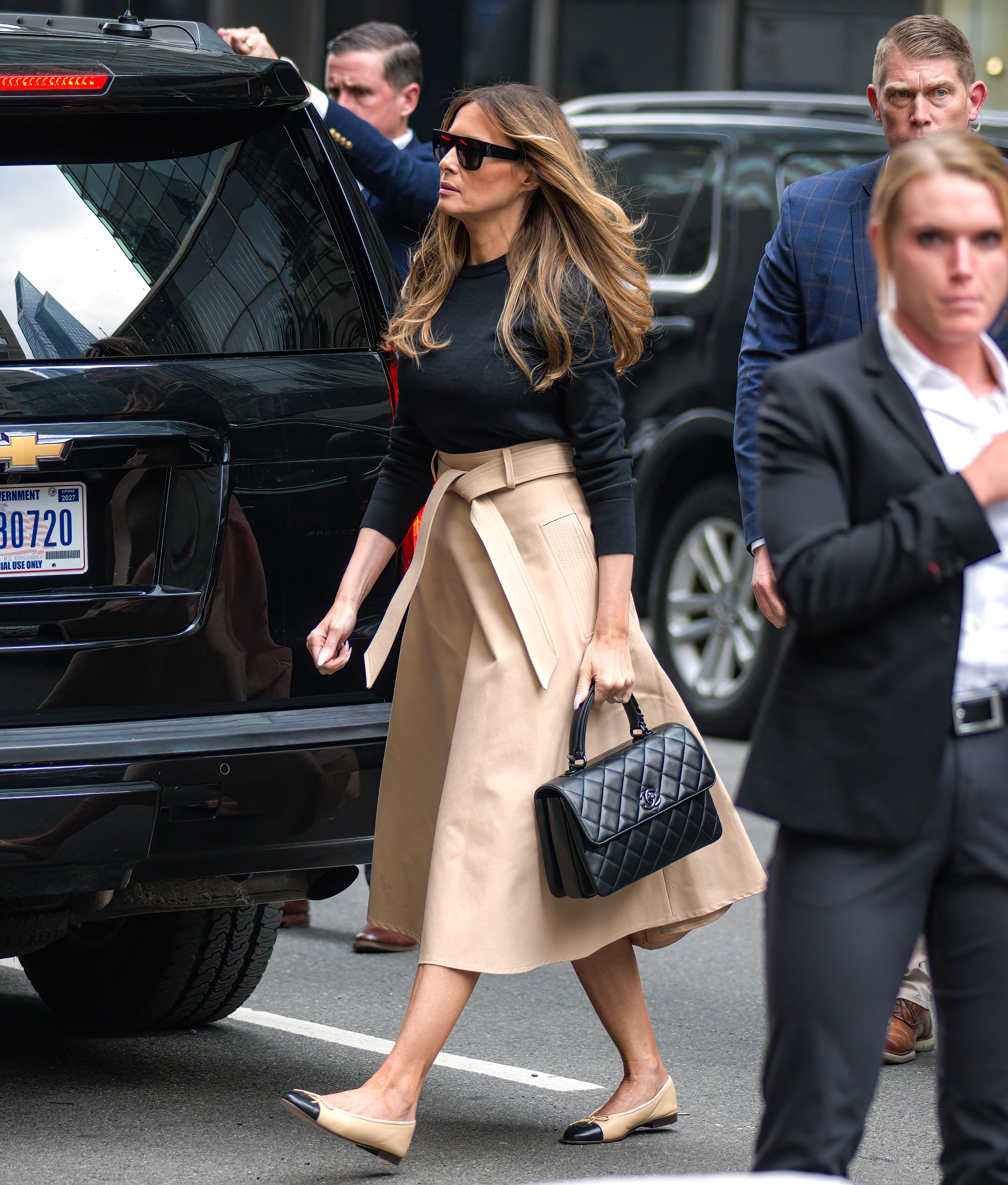 Melania Trump arrives at Trump Tower in New York City on June 8, 2023, wearing a black crewneck knit tucked into a high-waisted, tie-belt beige midi skirt with oversized sunglasses, pairing the polished street ensemble with a Chanel quilted handbag and classic two-tone beige-and-black cap-toe ballet flats.