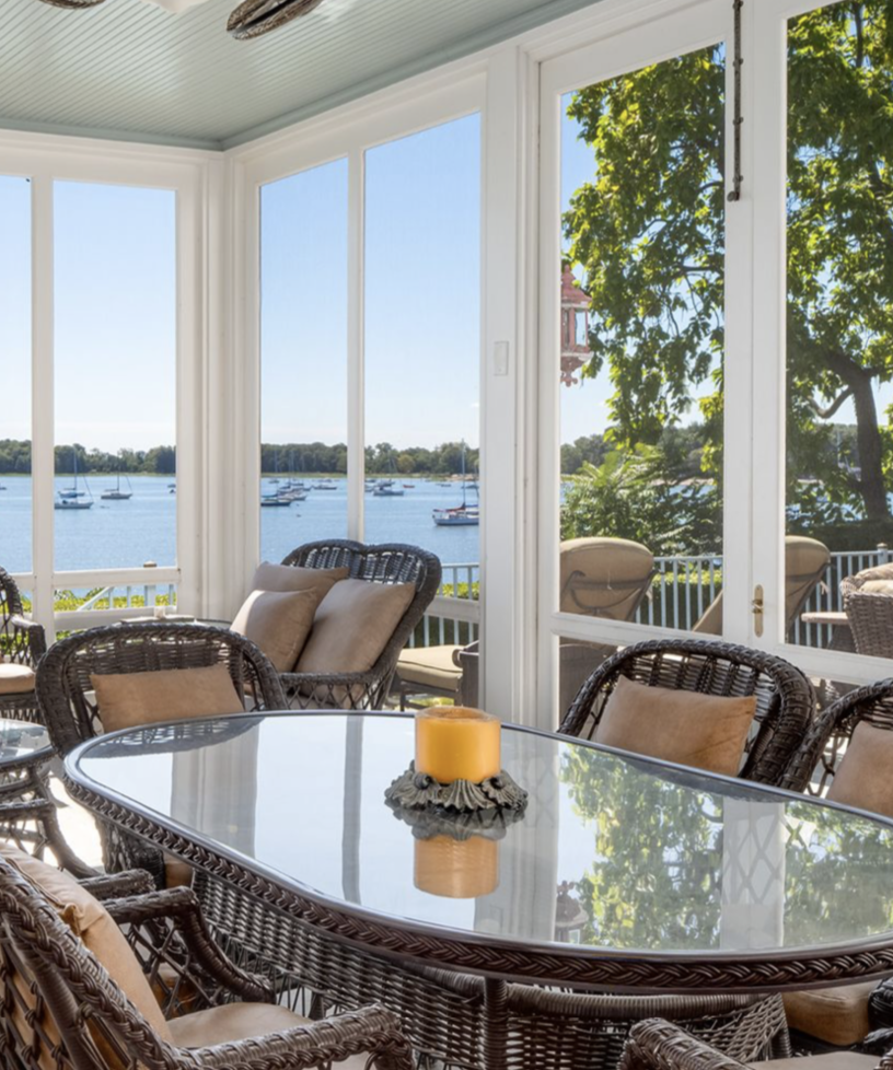 This sunlit sitting area at Cedar Cliff features wicker furniture, a glass-topped table, and floor-to-ceiling windows overlooking Long Island Sound — a peaceful spot to relax while enjoying the waterfront views. | Source: Instagram/@robbreport