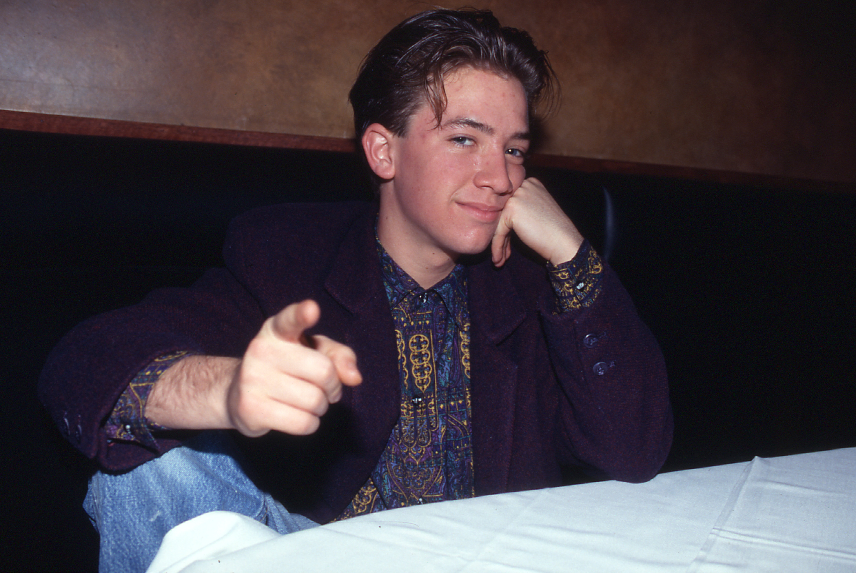 David Faustino flashed a charming smirk and pointed directly at the camera while seated at a restaurant booth in 1991. The "Married... with Children" star leaned his head on one hand, wearing a dark blazer layered over a vibrant, ornate paisley-patterned shirt and denim jeans. | Source: Getty Images