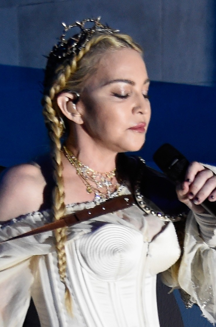 Madonna performs with long blond braids, layered necklaces and a white corset-style costume, holding a microphone as she looks downward. | Source: Chat GPT
