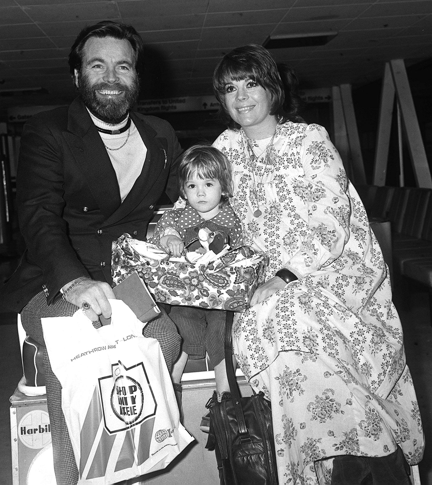 Robert Wagner and Natalie Wood sit with their three-year-old daughter Natasha Gregson Wagner at an airport in February 1974. Robert holds a duty-free shopping bag while Natalie, in a floral dress, smiles beside him. Natasha sits between them on a suitcase, holding a patterned travel bag with a toy inside.