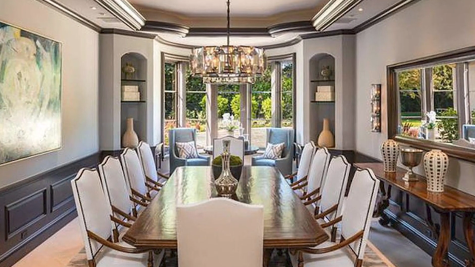 The formal dining room features a long polished-wood trestle table surrounded by high-back upholstered dining chairs, accented by dark wainscoting, arched display niches, an ornate chandelier, and a wide curved window bay overlooking the garden. | Source: YouTube/Taste of Country
