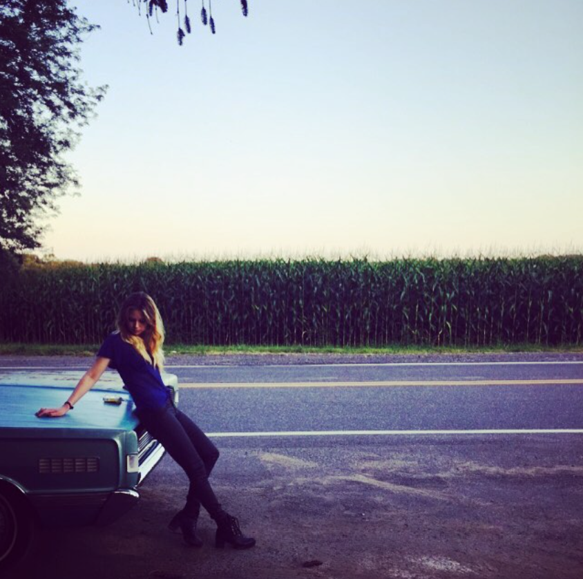 Daveigh Chase leans against a parked vehicle beside a rural road bordered by tall crops in a photo posted in August 2015, during the years before she largely disappeared from public view. | Source: Instagram/daveighc