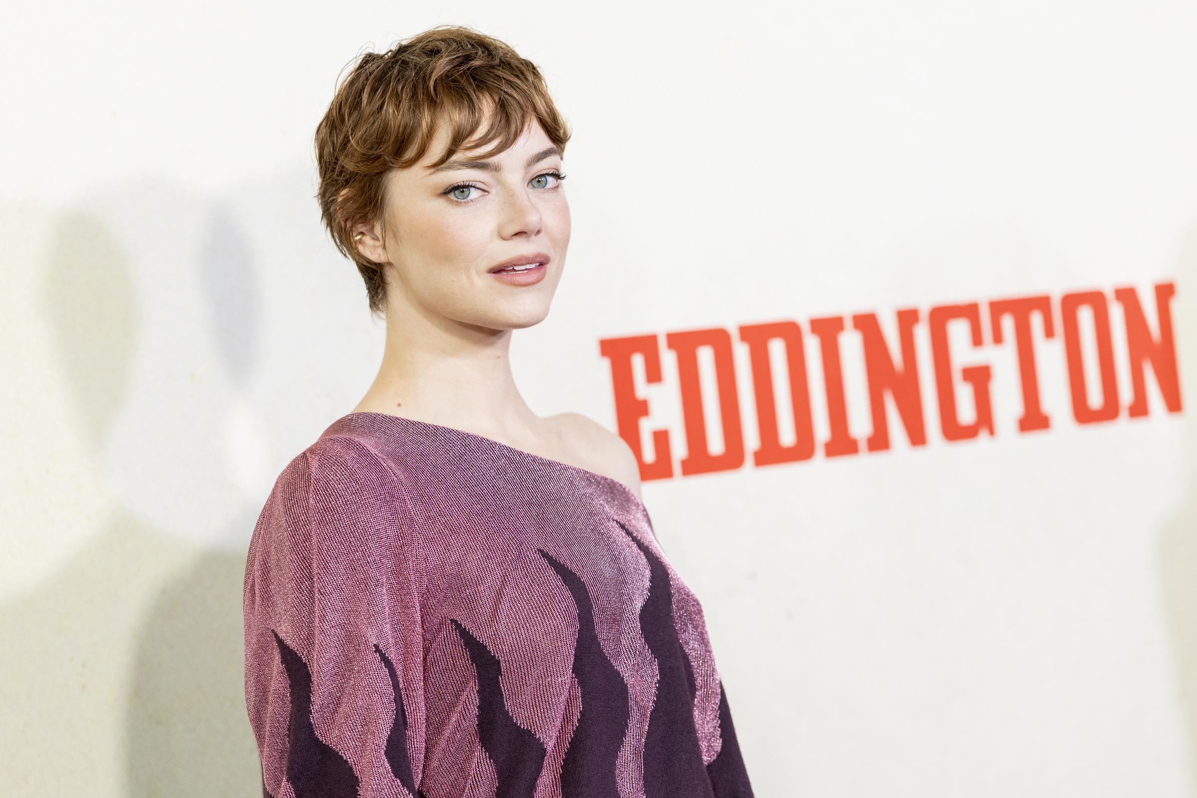 With a confident and magnetic gaze, Stone embodies effortless poise, a subtle smirk playing on her lips. Her striking presence, accentuated by the hint of her shimmering eggplant one-shoulder dress, takes center stage against the bold, dark orange "Eddington" lettering in the background.