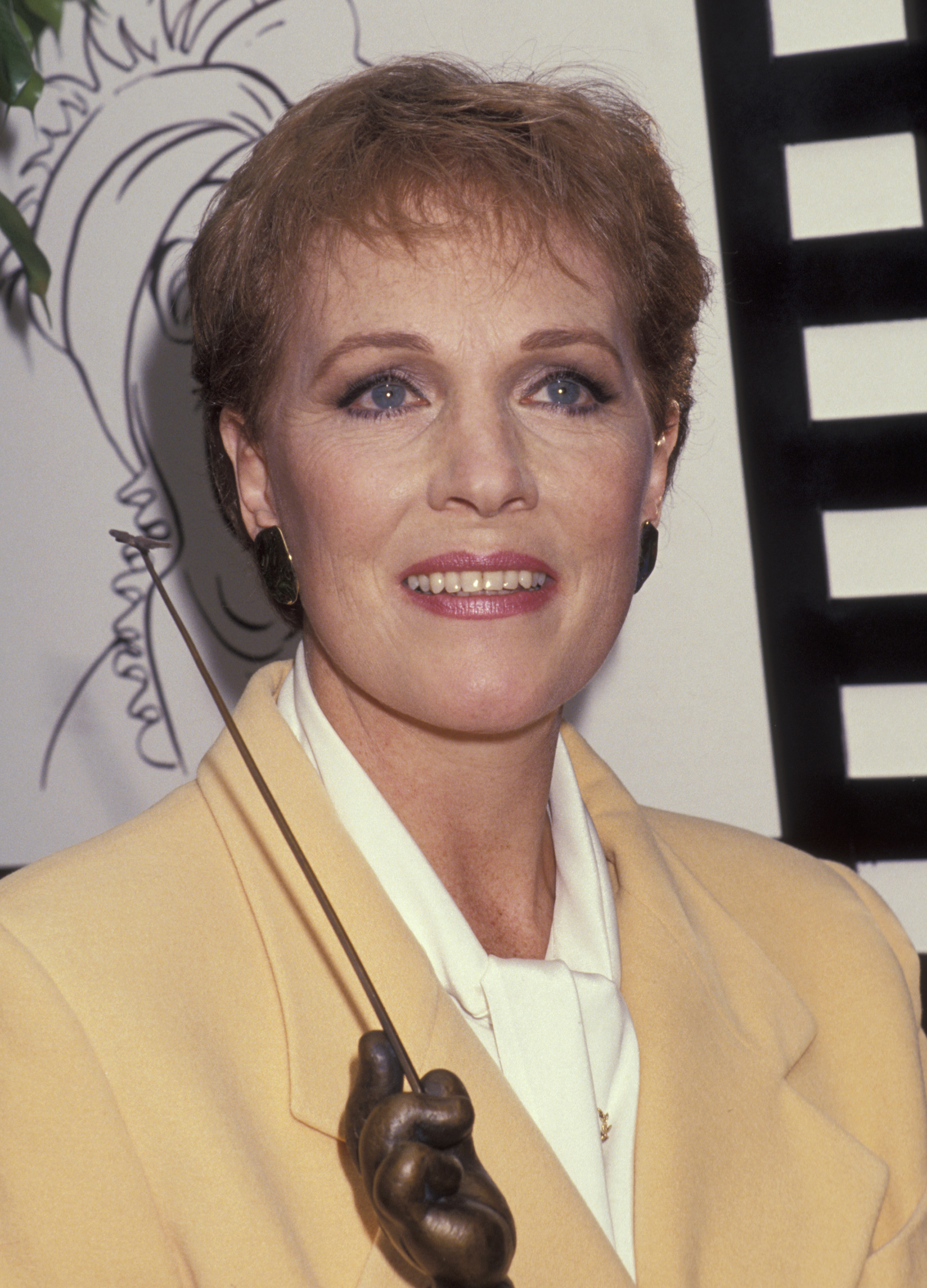 Julie Andrews beams while holding her bronze trophy during the Disney Legends Awards ceremony at Disney Studios in Burbank, California, on October 22, 1991. Honored for her indelible cinematic legacy with the studio, the actress looks luminous in a soft pastel yellow blazer layered over a crisp ivory ascot blouse, paired with dark statement clip-on earrings and her signature cropped haircut.