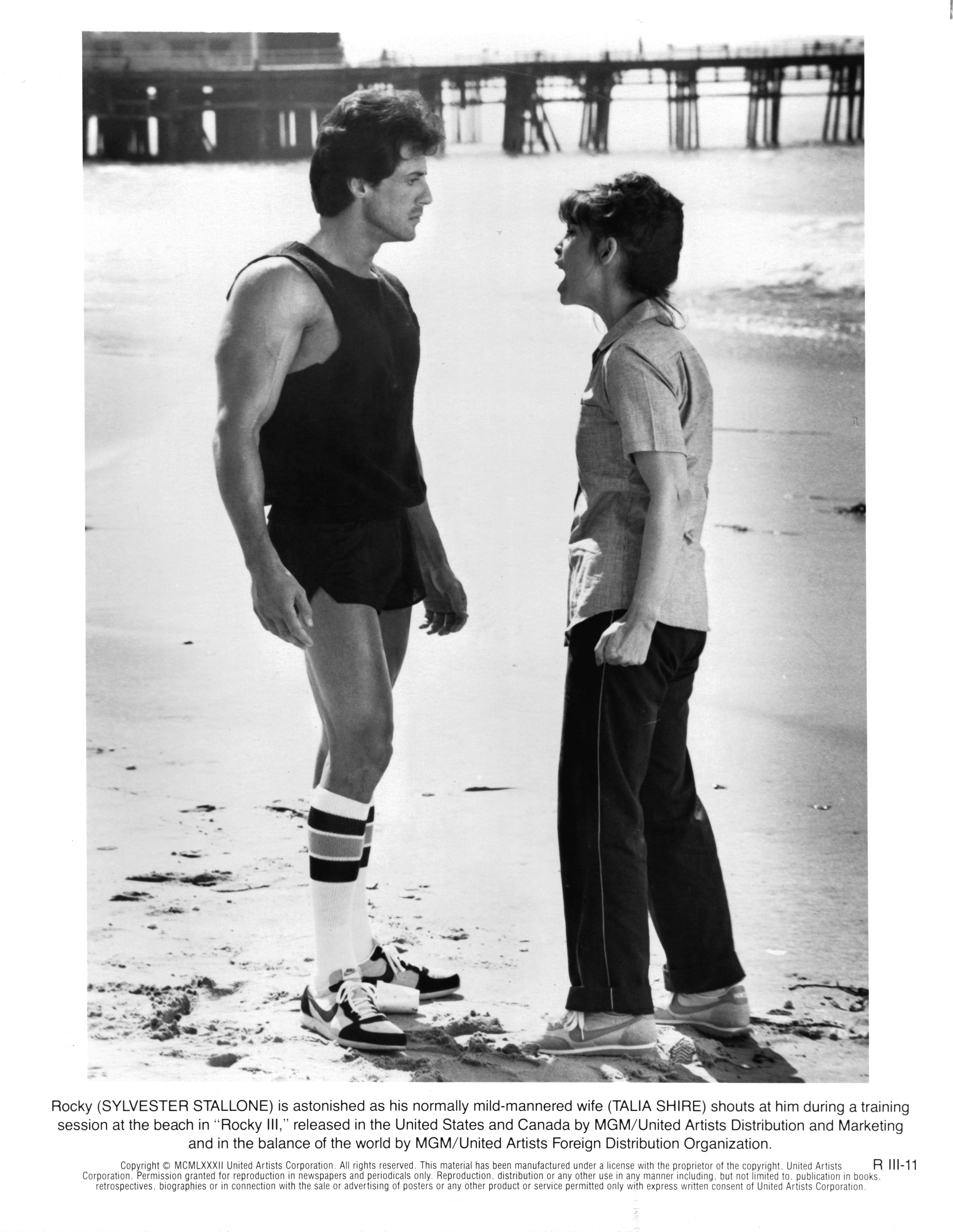 Sylvester Stallone faces Talia Shire during a beach training scene from "Rocky III" in 1982, with Shire's Adrian appearing to confront Rocky as he prepares for his next fight. Dressed in training gear, Stallone's noticeably lean frame reflects the physical transformation he underwent to give Rocky greater speed and agility against Clubber Lang.