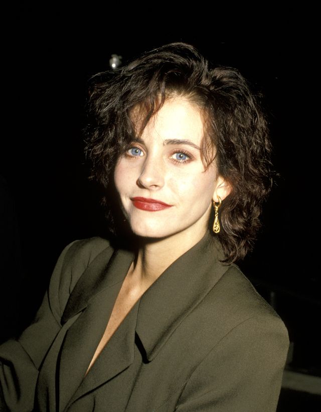 Courteney Cox stuns in a 1990 close-up, highlighting her striking blue eyes, bold red lipstick, and gold dangling earrings, all paired with a stylish green suit.