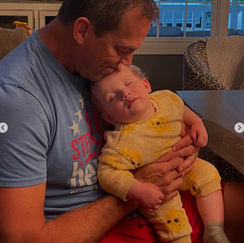Bryon Noem cradles a sleeping child in his arms, gently resting his head against the child’s forehead in a quiet indoor setting. | Source: Instagram/kristinoem
