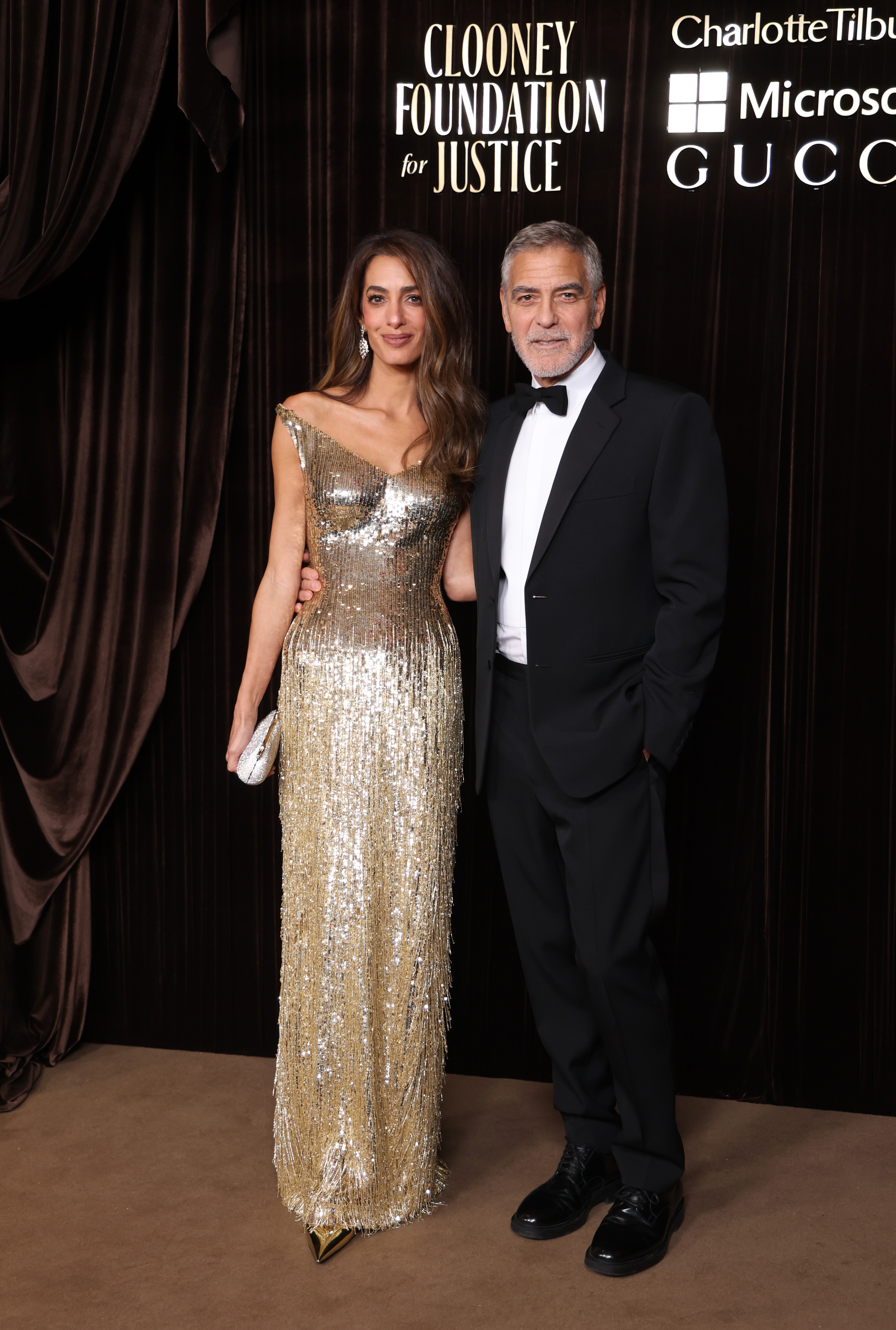 touches several generations of the family. George Clooney and his wife, Amal Clooney, pose together at The Albies, the annual awards ceremony hosted by the Clooney Foundation for Justice. Amal wears a shimmering gold, floor-length gown with an asymmetrical neckline and matching dangling earrings, while George looks sharp in a black tuxedo, white shirt, and bow tie. | Source: Getty Images