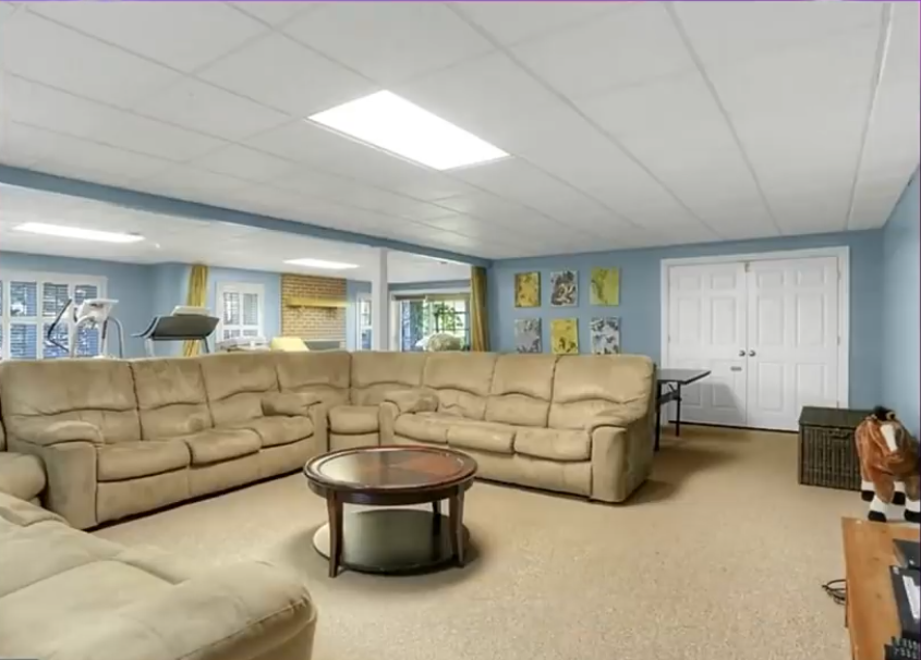 The basement family room features a large beige sectional sofa arranged around a round wooden coffee table. Pale blue walls, neutral carpeting, a drop ceiling, and recessed lighting complete the spacious area, with exercise equipment visible in the background. | Source: YouTube/Without A Crystal Ball