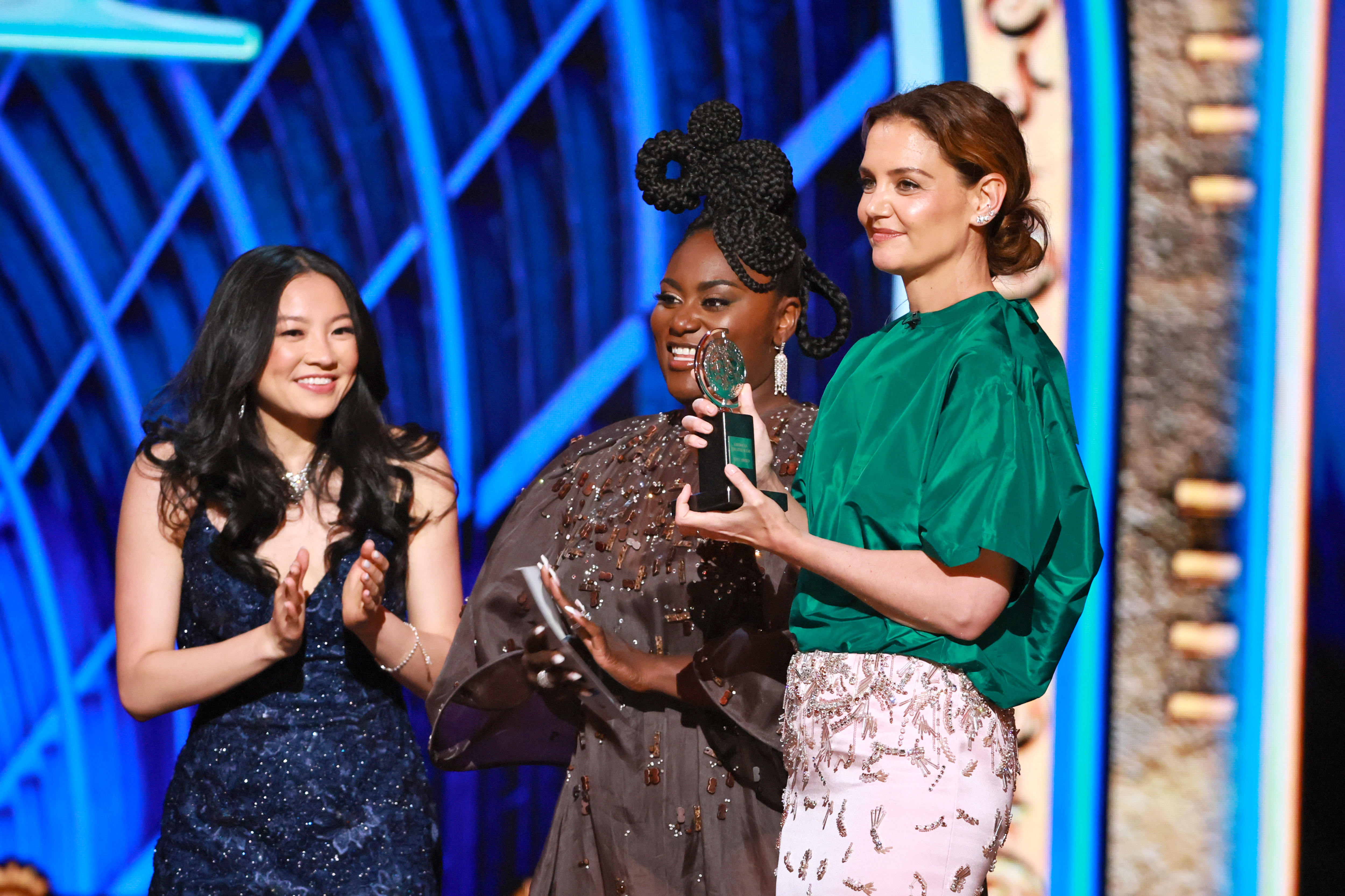 Katie Holmes joins Danielle Brooks and a smiling honoree onstage, celebrating a Tony Award win. The moment reflects both style and joy, with Holmes in her standout ensemble applauding the night’s talent.