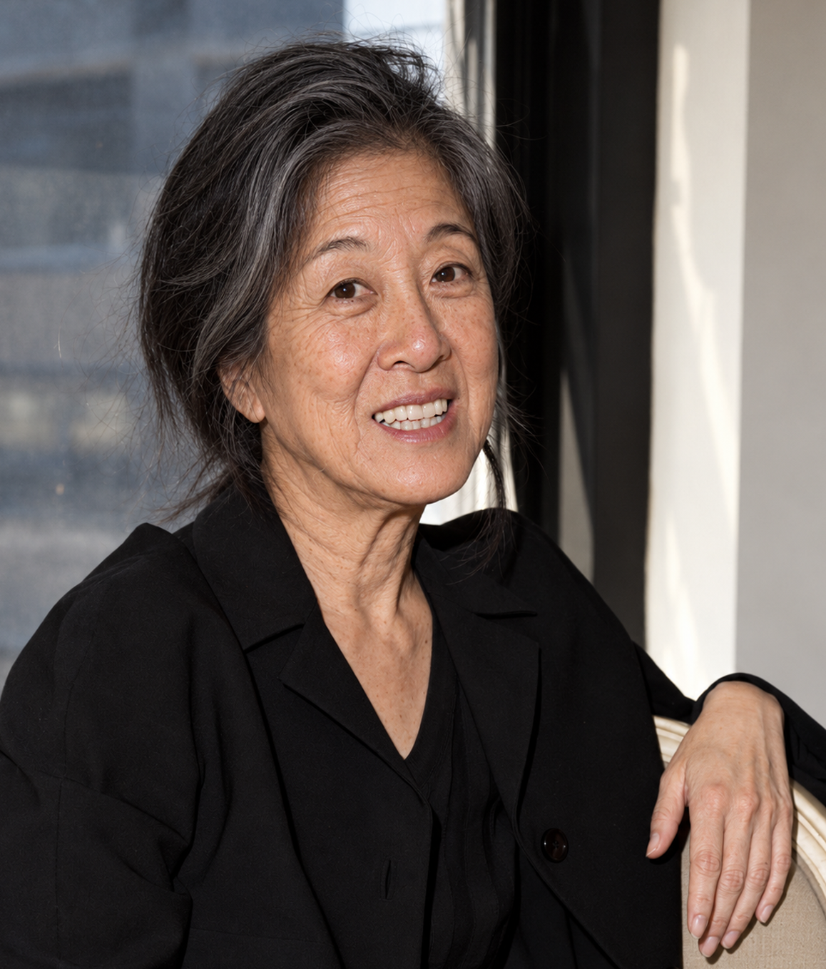An AI-generated portrait reimagines Vera Wang aging gracefully with silver-streaked hair, soft laugh lines, and her original, unaugmented facial features. Resting against the back of an armchair by a sunlit window, the conceptual visual depicts the bridal designer smiling warmly while dressed in a classic black button-up blazer. | Source: ChatGPT